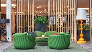 Eine moderne Lobby mit grünen Sitzgelegenheiten, einem schwarzen Pflanzgefäß mit Pflanzen und einer gelben Lampe.