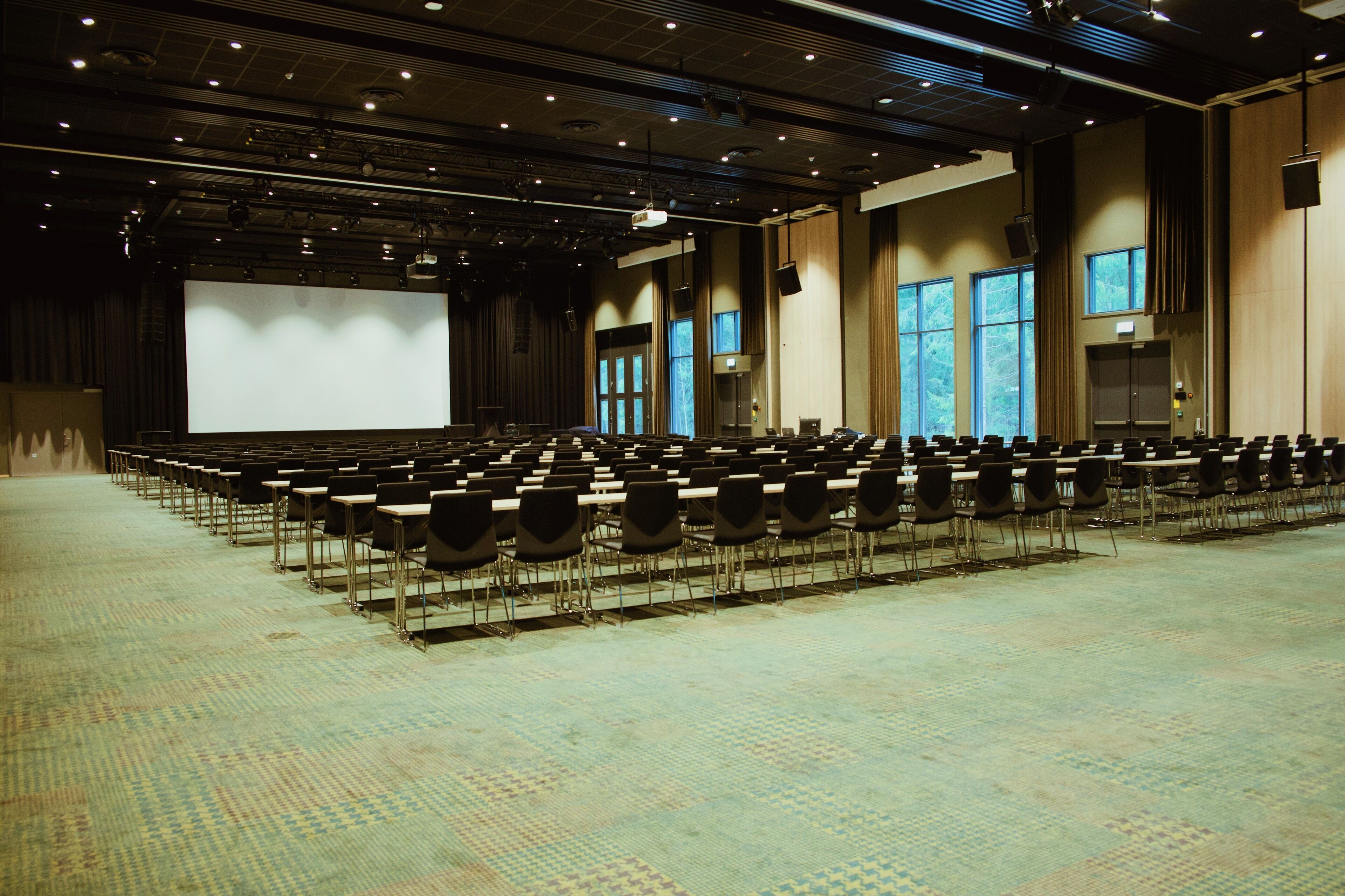 A large conference room with rows of chairs, a stage with a large screen, and windows providing natural light.
