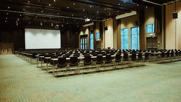 A large conference room with rows of chairs, a stage with a large screen, and windows providing natural light.