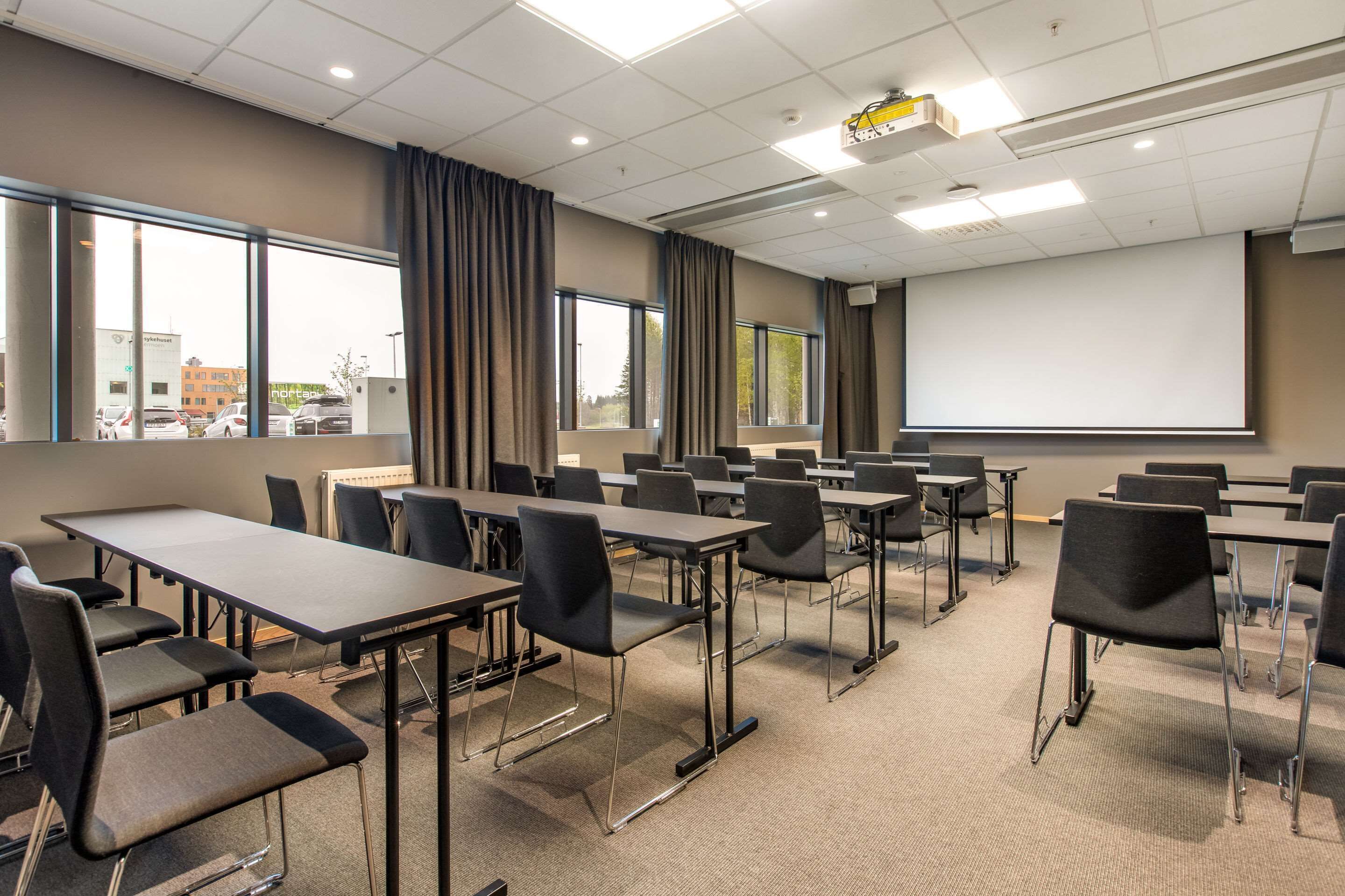 A conference room with tables and chairs, large windows, a projector screen, and a view of buildings outside.