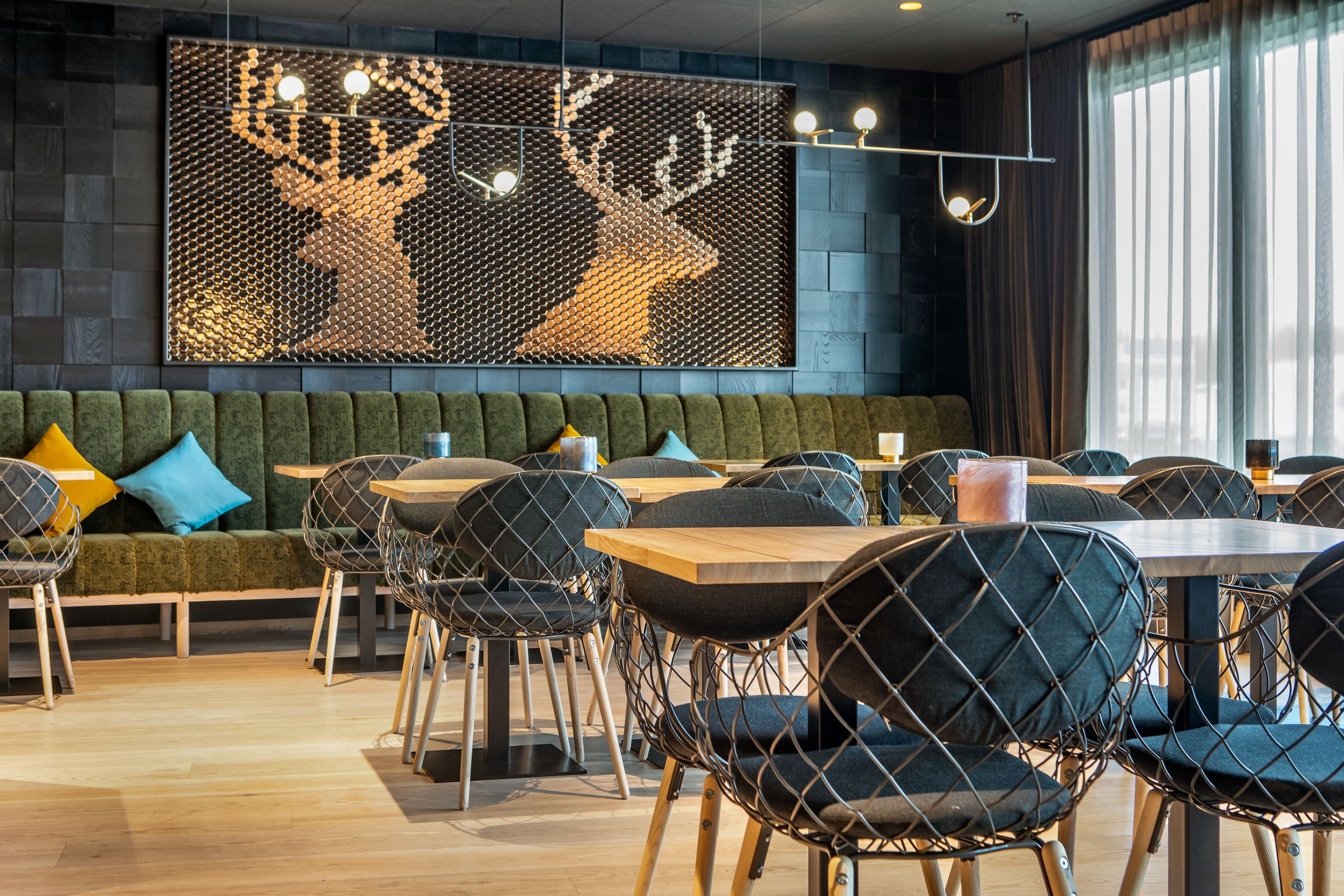 Um restaurante moderno com um banco acolchoado verde, cadeiras de estrutura de arame preto e uma grande obra de arte de veados na parede.