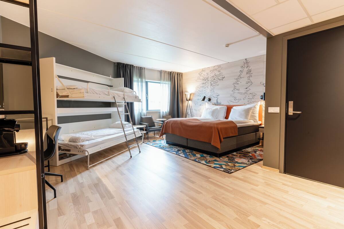 Een moderne hotelkamer met een stapelbed, een bureau met stoel, een klein tafeltje en een bed met een kamerkleed met patroon.