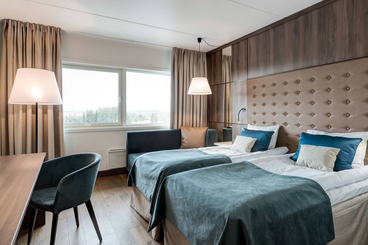 Een hotelkamer met twee bedden, een bureau en een stoel. Er is een raam met gordijnen, een lamp en een wand met een gestoffeerd hoofdeinde.