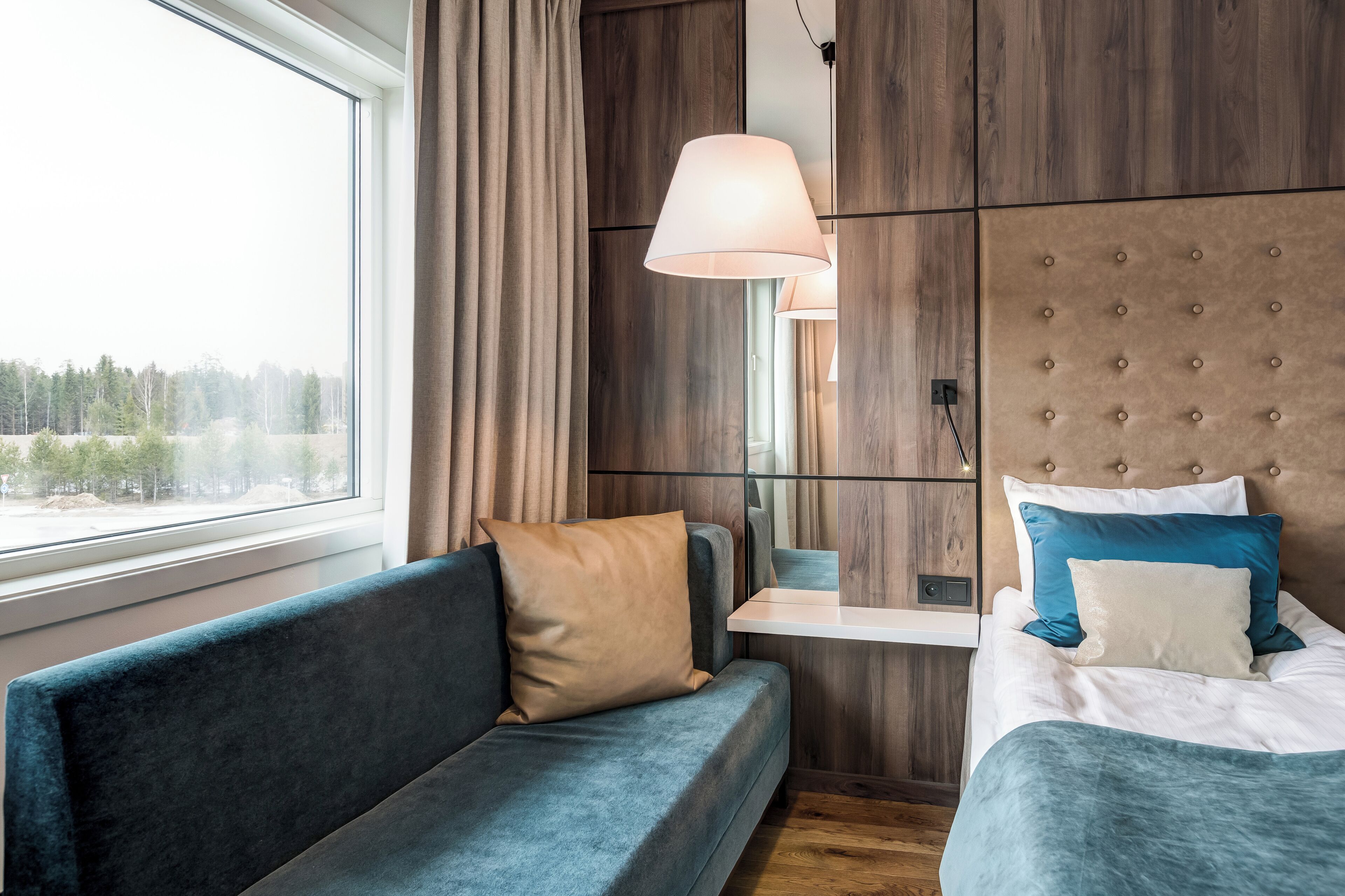 A modern hotel room with a sofa, a bed with blue and white bedding, a large window with curtains, and a wooden headboard.