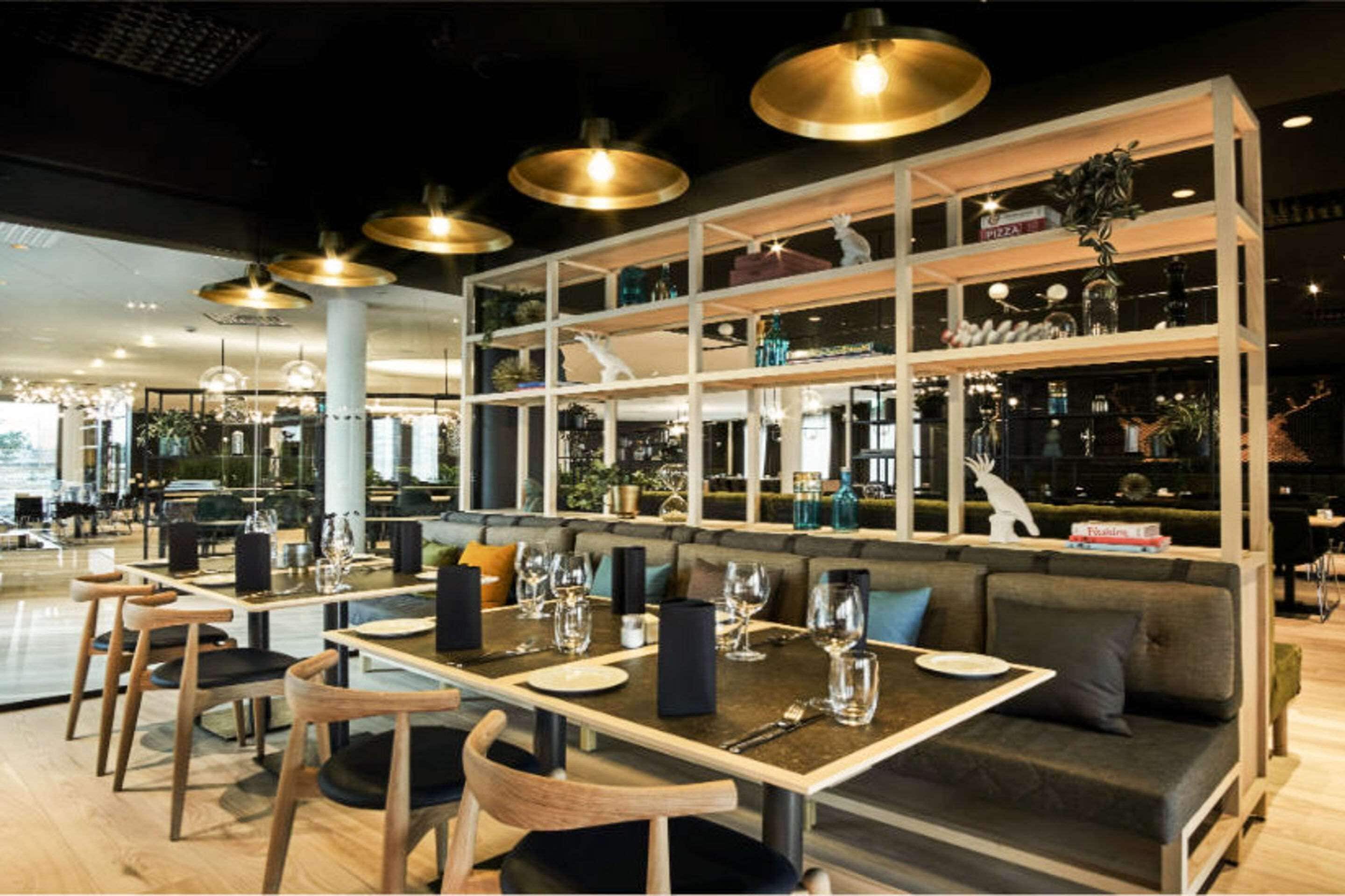Un interior de restaurante moderno con mesas de madera, asientos acolchados, iluminación colgante y varios objetos decorativos en estantes.
