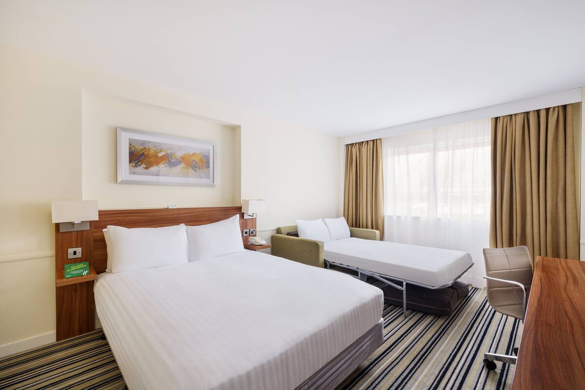A hotel room with two single beds, a desk, a chair, a nightstand with a lamp, and a painting on the wall.