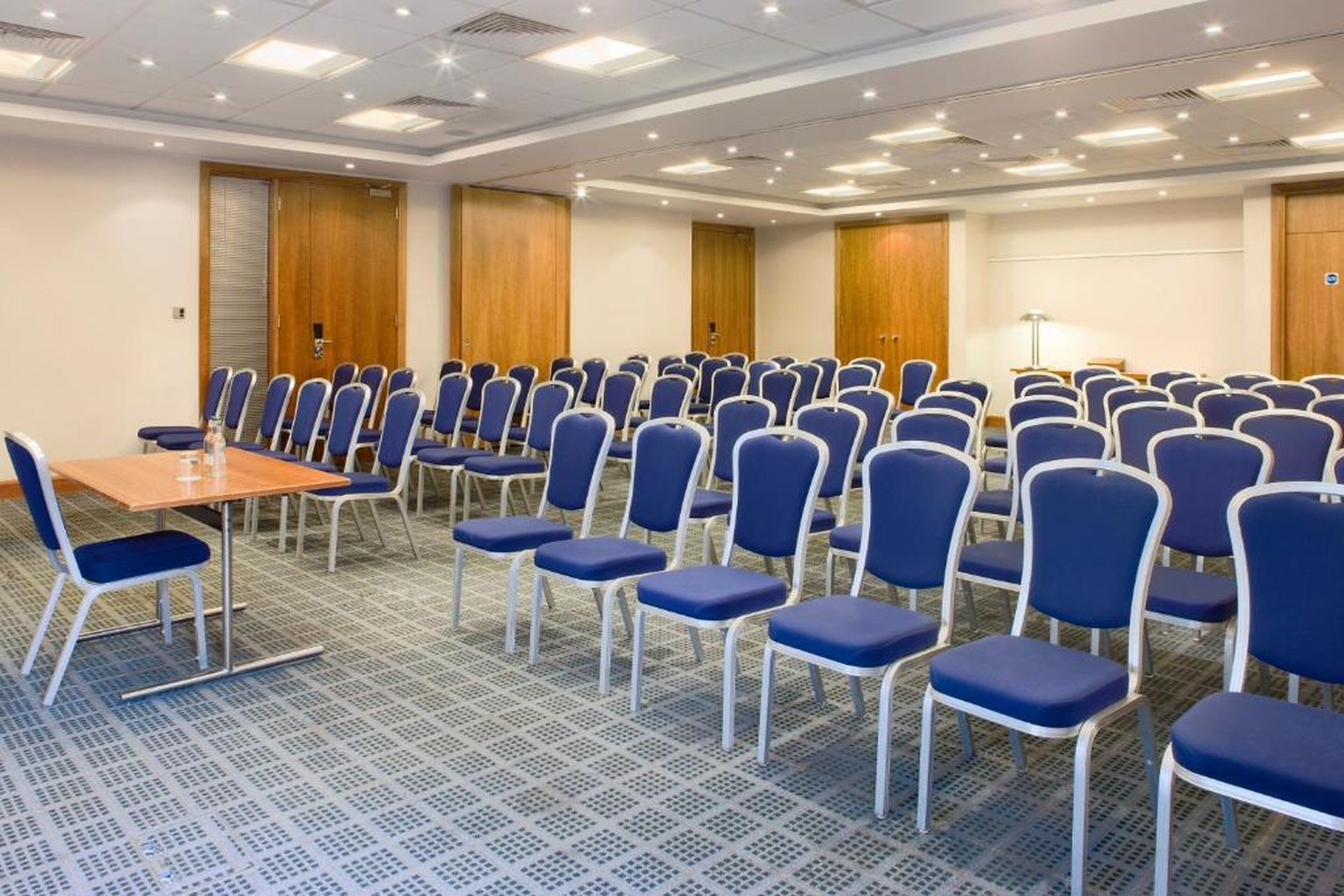 Un salón de conferencias con filas de sillas azules, una mesa de madera y una pantalla de proyector.
