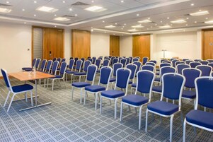 Un salón de conferencias con filas de sillas azules, una mesa de madera y una pantalla de proyector.