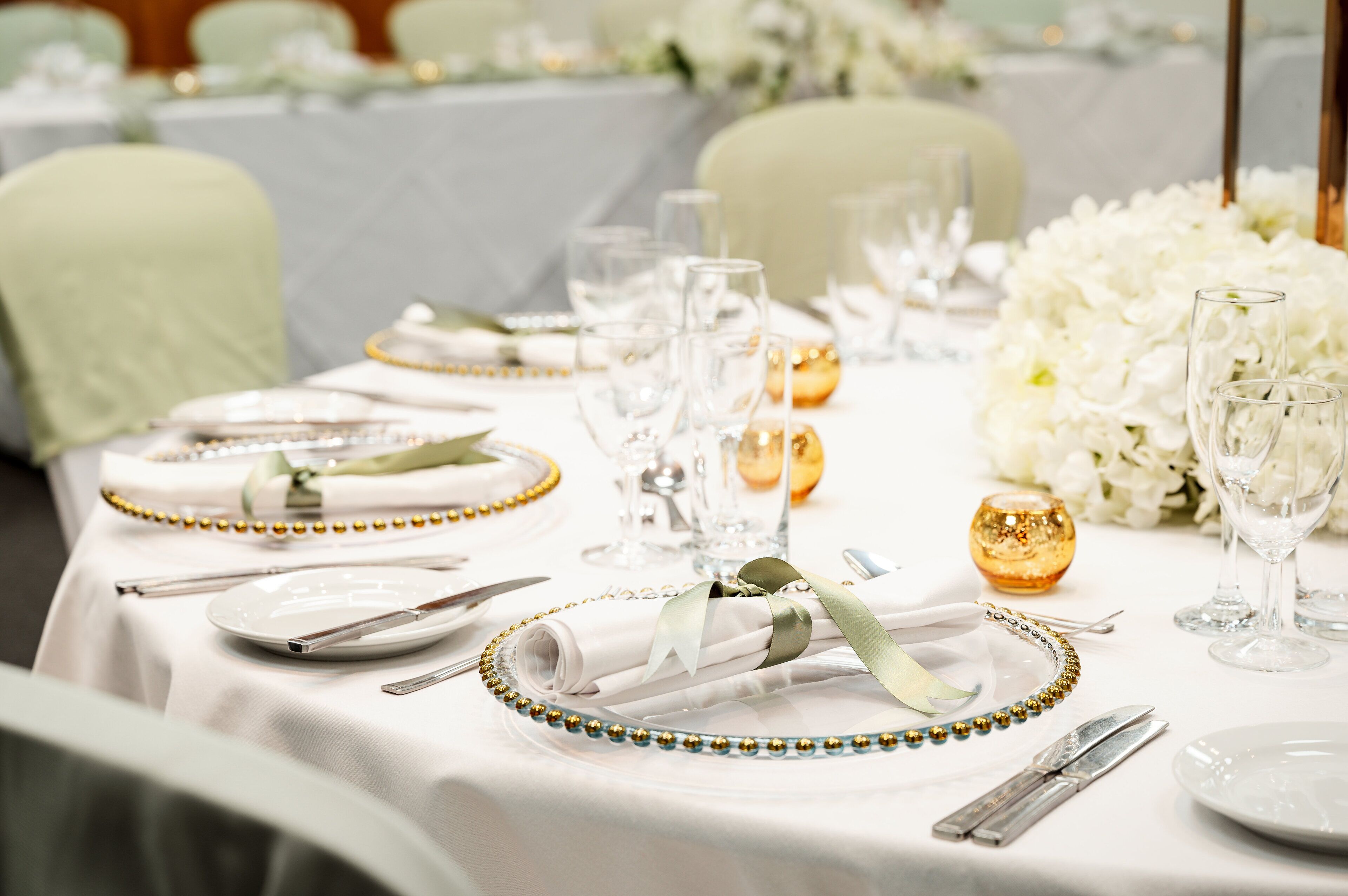 A table set for a formal event with white tablecloths, gold and green decorative elements, and white floral arrangements.