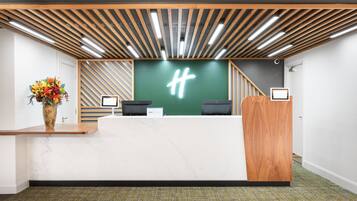 Un mostrador de recepción con paneles de madera, una pared verde con un logotipo blanco y un jarrón con flores.