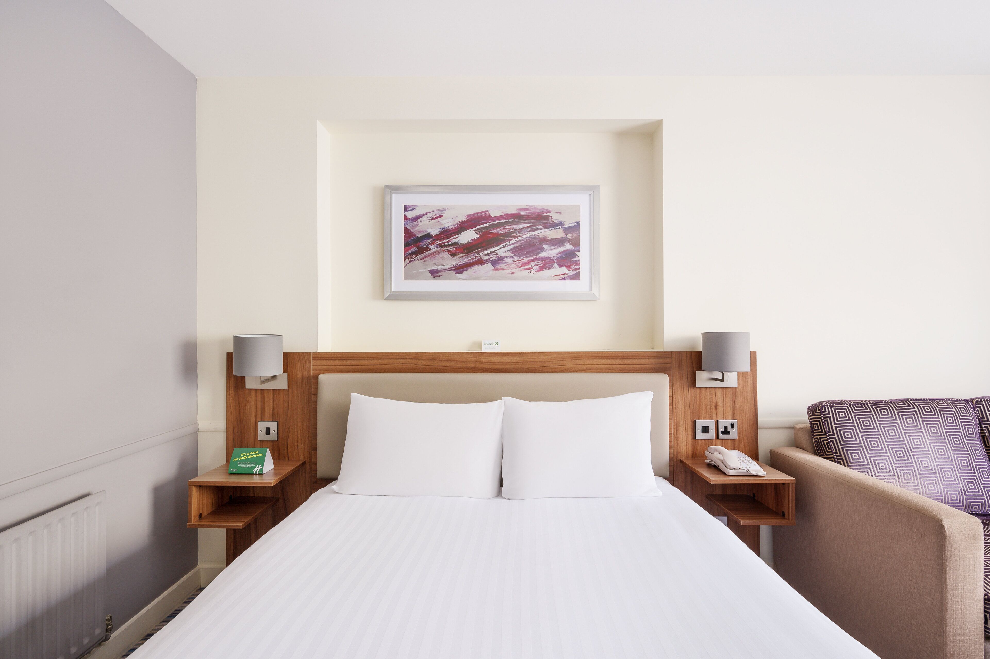 Habitación de hotel con una cama, cabecero, un cuadro abstracto enmarco, lámparas de noche, un teléfono y un pequeño estante con un libro.