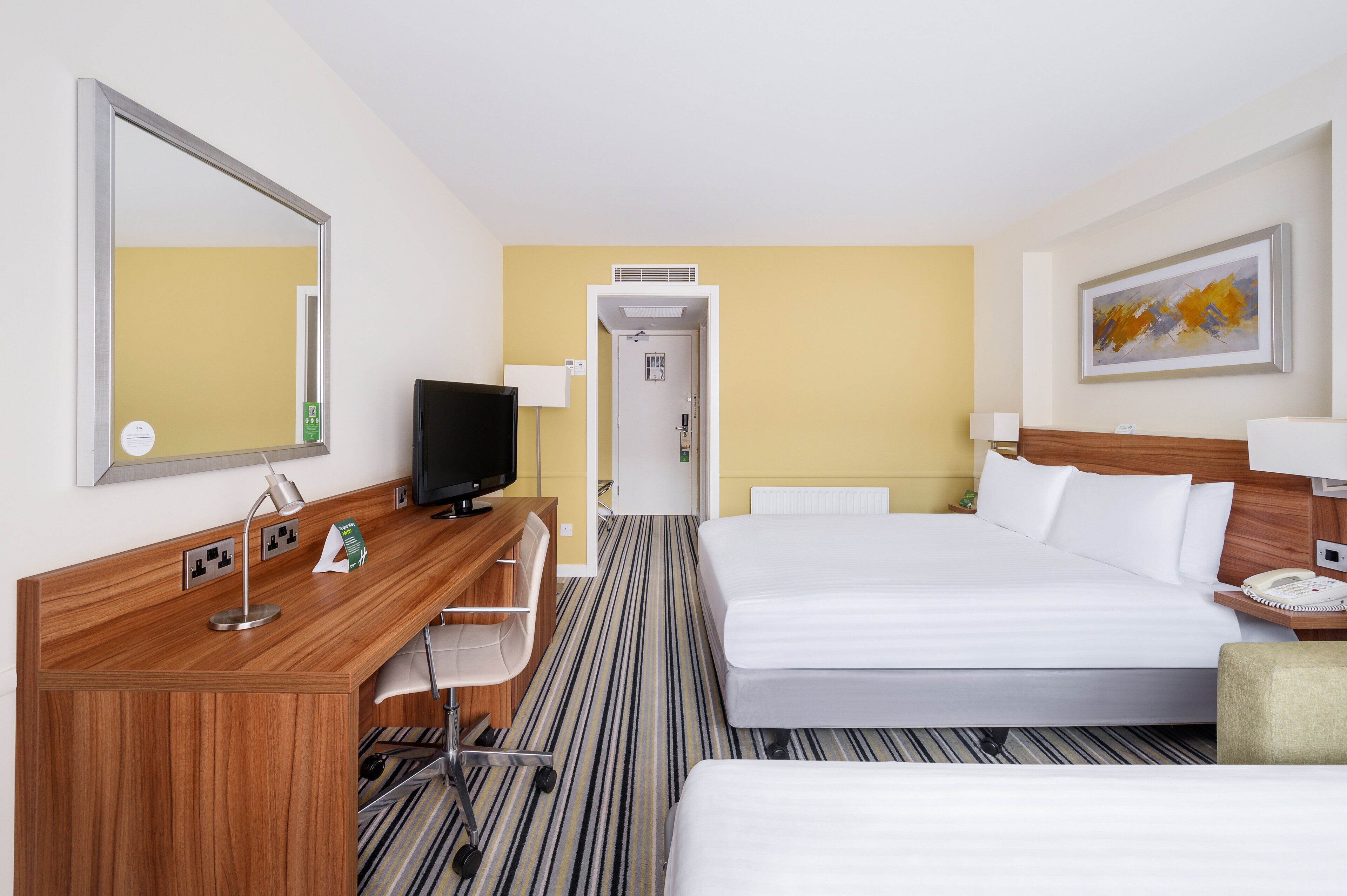 Quarto de hotel com escrivaninha de madeira, televisão, cama e mesa de cabeceira com abajur.