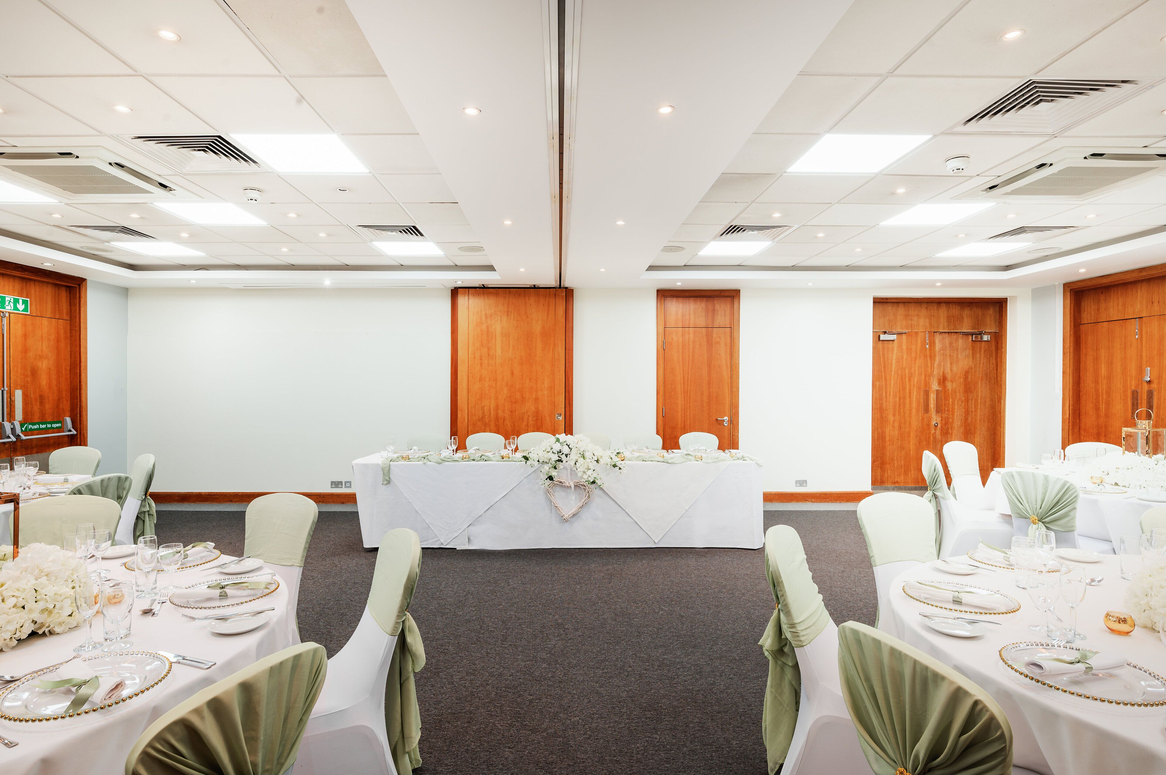 A conference room with a long table set for a meeting, surrounded by chairs with green sashes, white tablecloths, and floral centerpieces.
