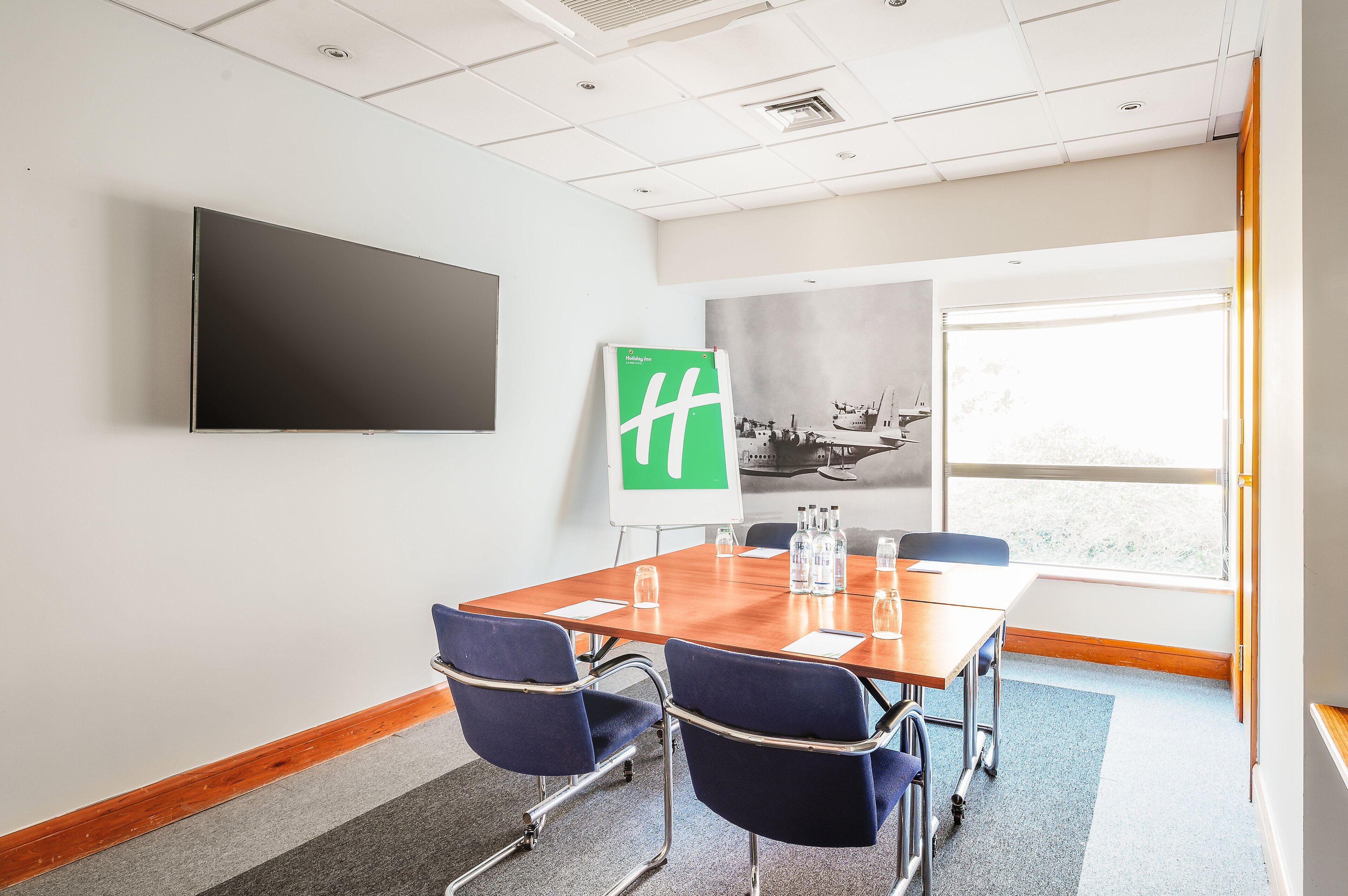 Un salón de conferencias con una mesa grande, cuatro sillas, un televisor de pantalla plana, una pizarra y una ventana con vistas al exterior.