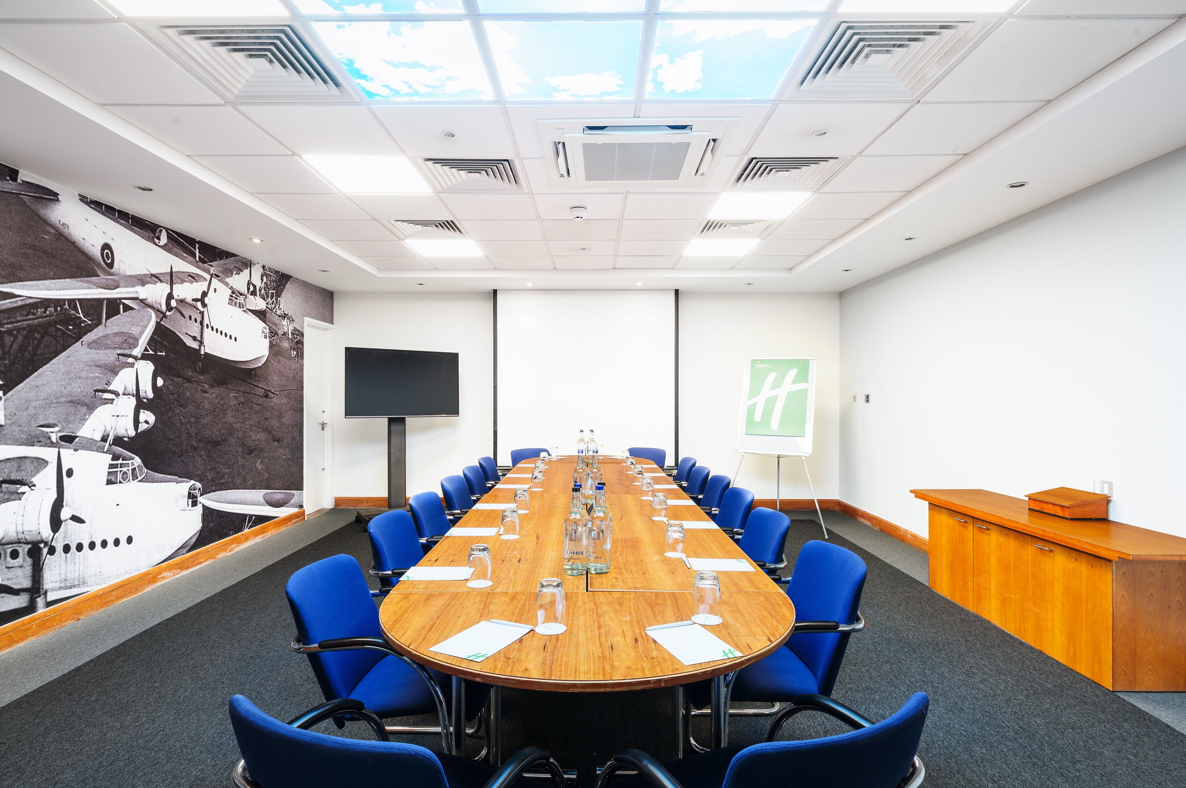 Ein Konferenzraum mit einem langen Tisch, blauen Stühlen, einer Projektionsleinwand und einem Wandgemälde mit Flugzeugen.