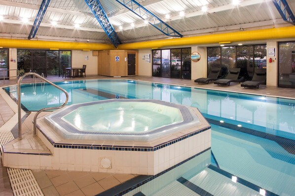 An indoor swimming pool with a hot tub, lounge chairs, and a covered roof structure.