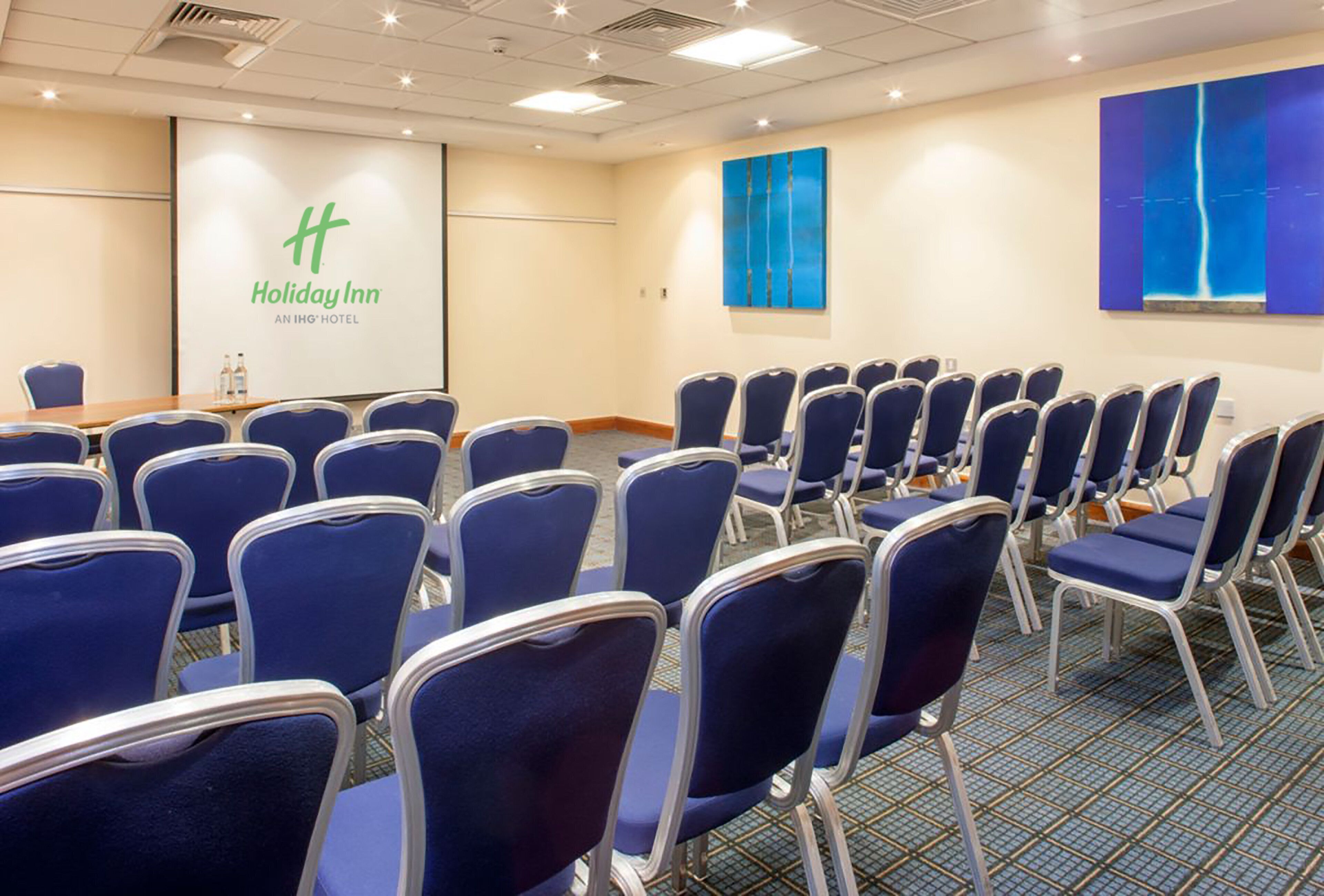 Un salón de conferencias con hileras de sillas azules, una pizarra blanca con la marca 'Holiday Inn' y dos cuadros abstractos en las paredes.