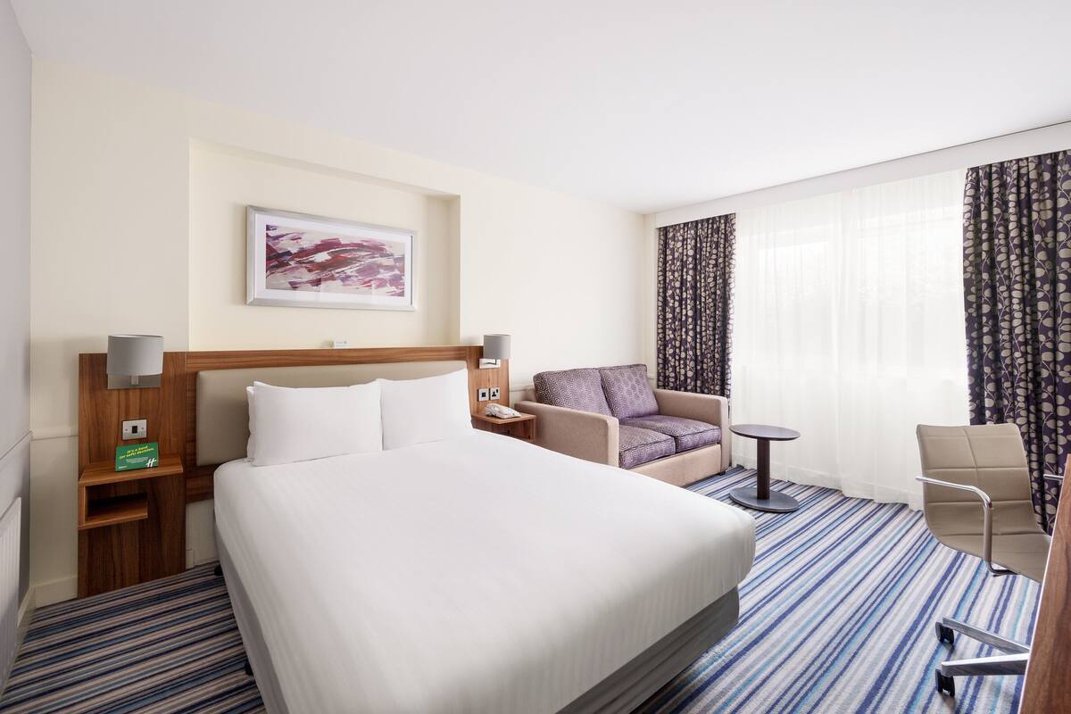 A hotel room with a large bed, a small sofa, a chair, a table, a lamp, and a painting on the wall.