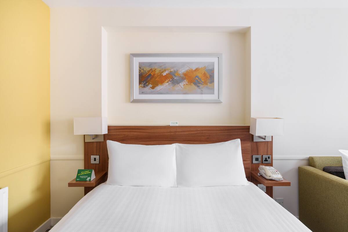 A hotel room with a bed, two bedside tables, a telephone, a lamp, and a painting on the wall.