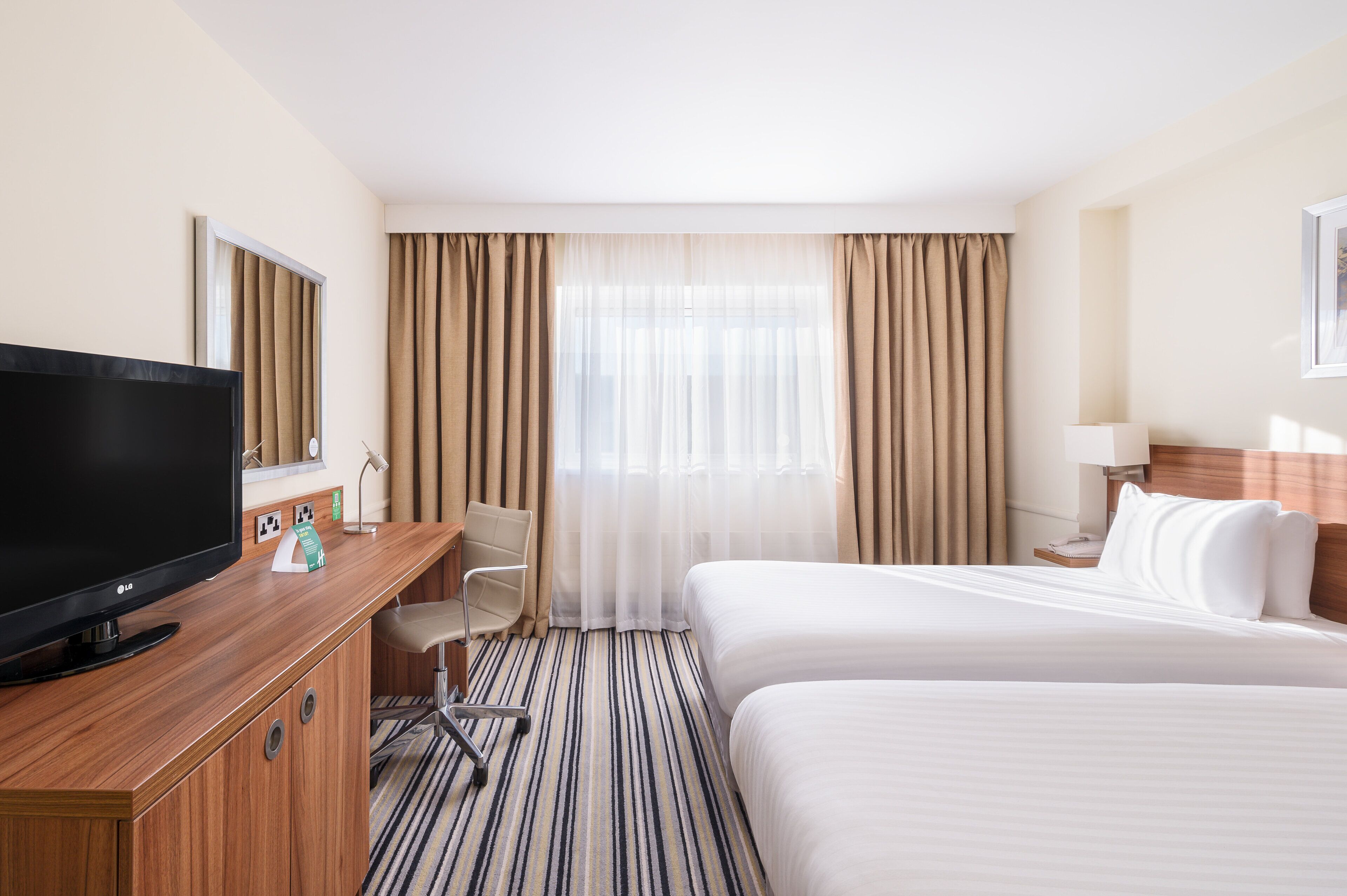 A hotel room with a large flat-screen TV, a desk with a chair, a bed with white linens, and a window with curtains.