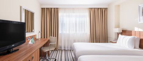 A hotel room with a large flat-screen TV, a desk with a chair, a bed with white linens, and a window with curtains.