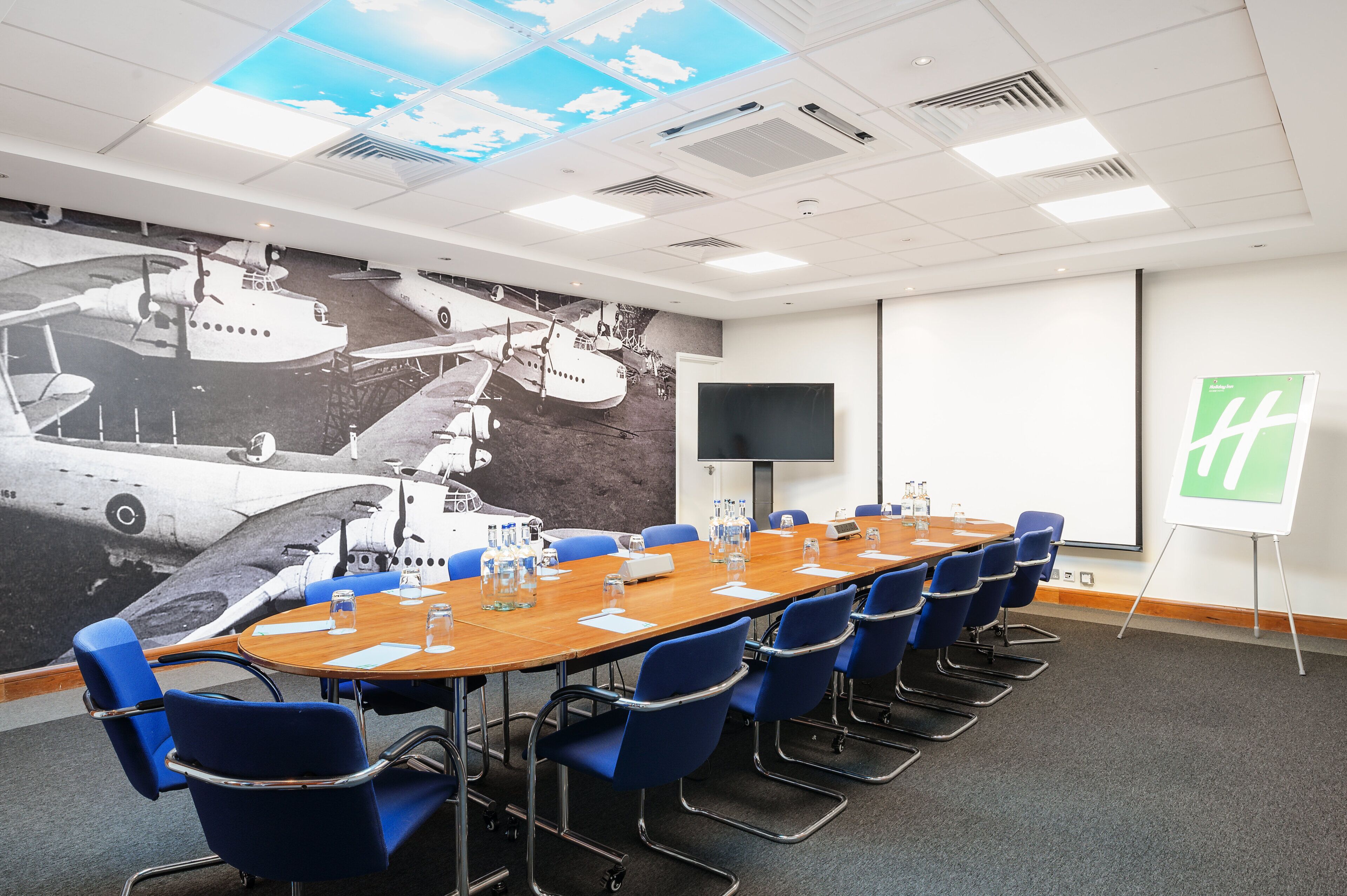 Un salón de conferencias con una larga mesa, sillas azules, una pantalla de proyector y un gran mural de un avión en la pared.