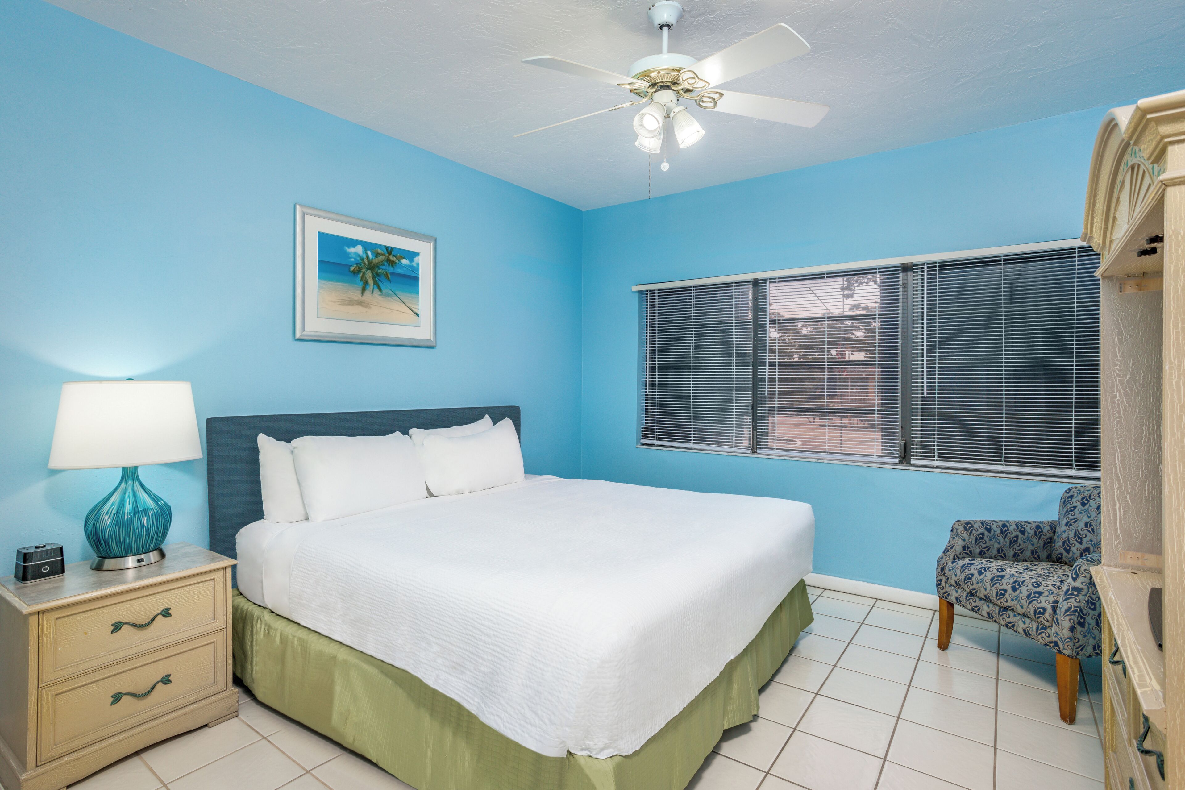 A bedroom with a bed, a nightstand, a chair, a ceiling fan, a window with blinds, and a framed picture on the wall.