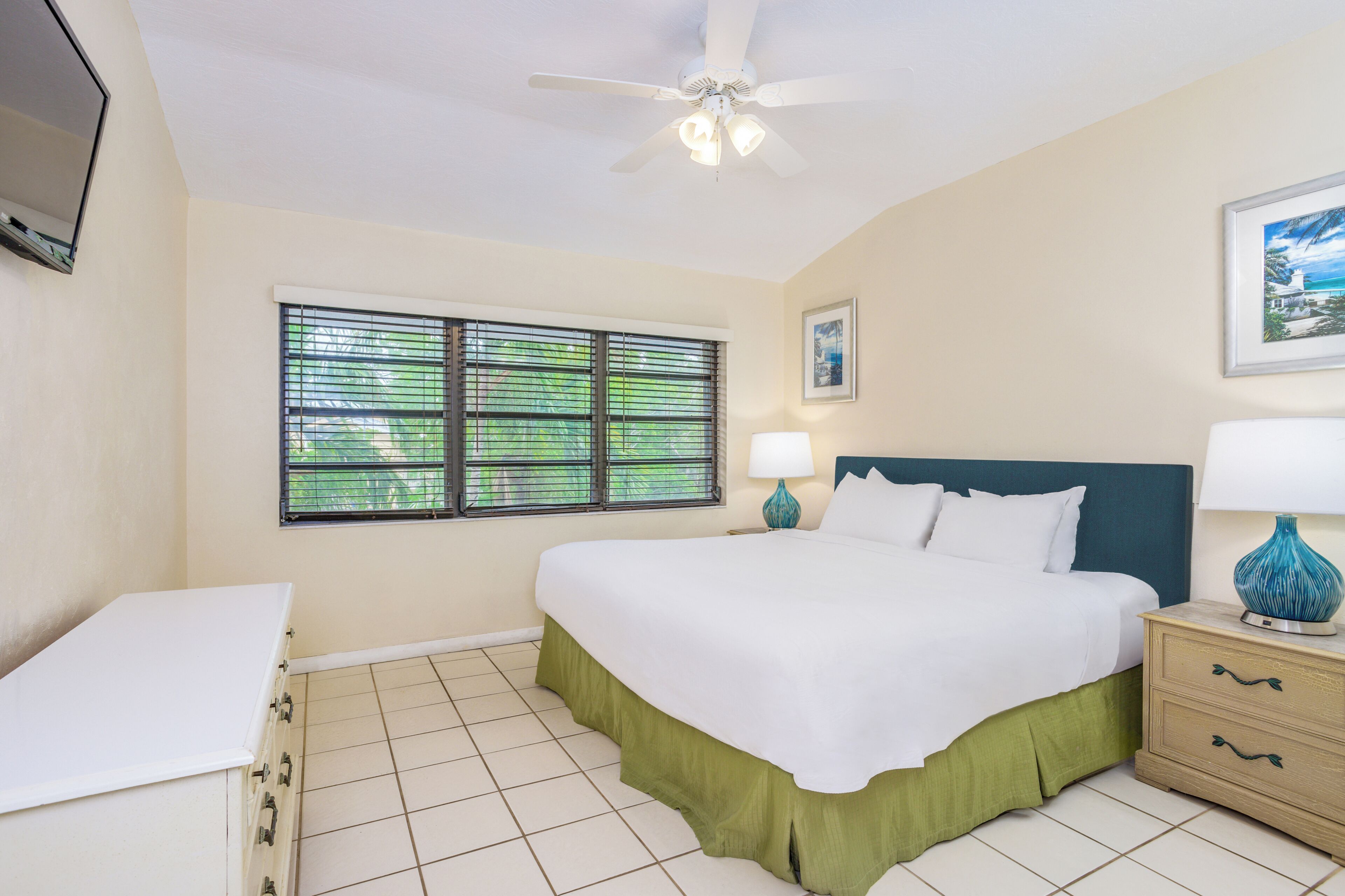 A hotel room with a bed, a nightstand, a lamp, a ceiling fan, a window with blinds, and a framed picture on the wall.