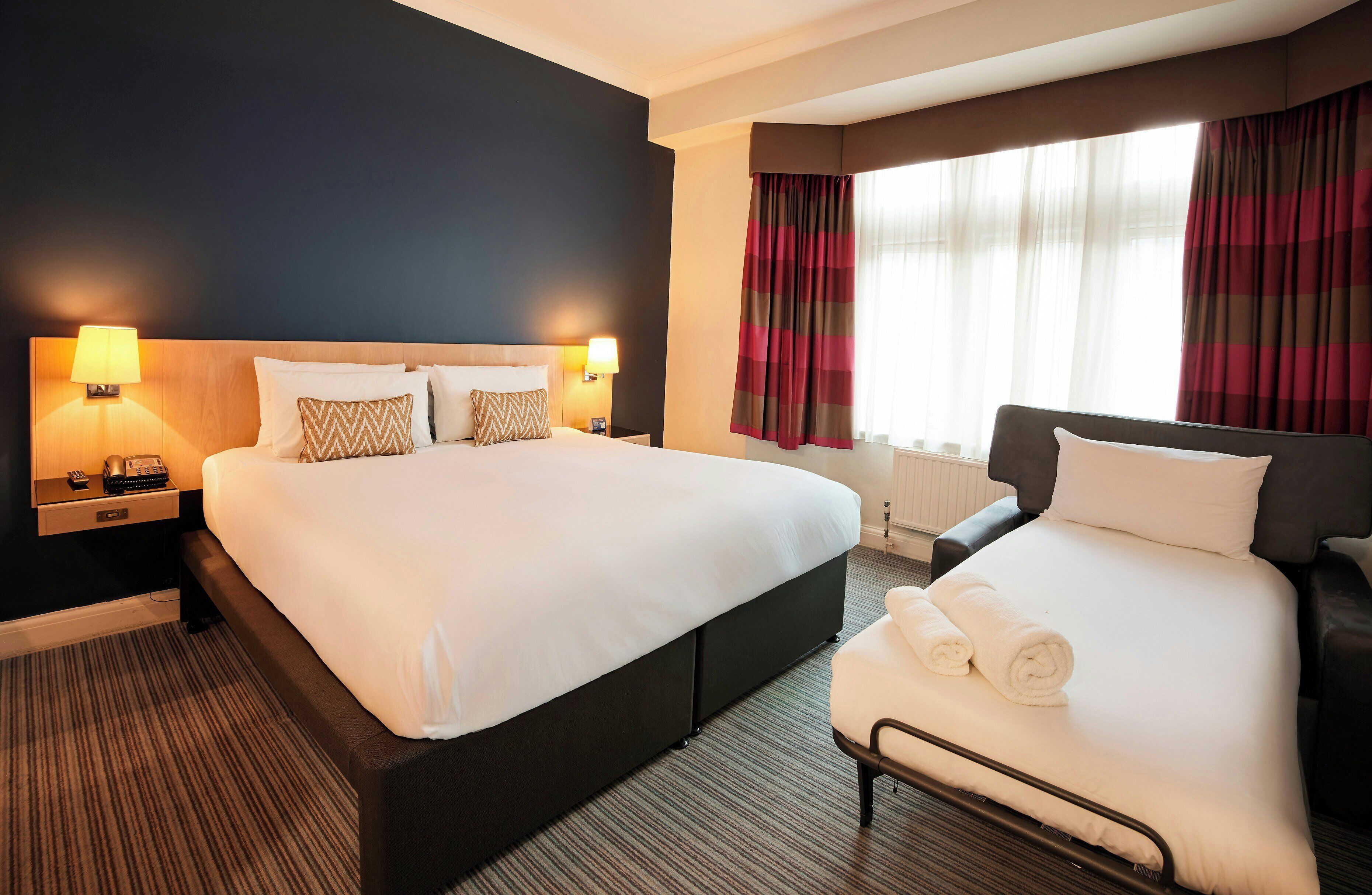 Habitación de hotel con una cama grande, una cama más pequeña, una mesita de noche con lámpara y una ventana con cortinas.