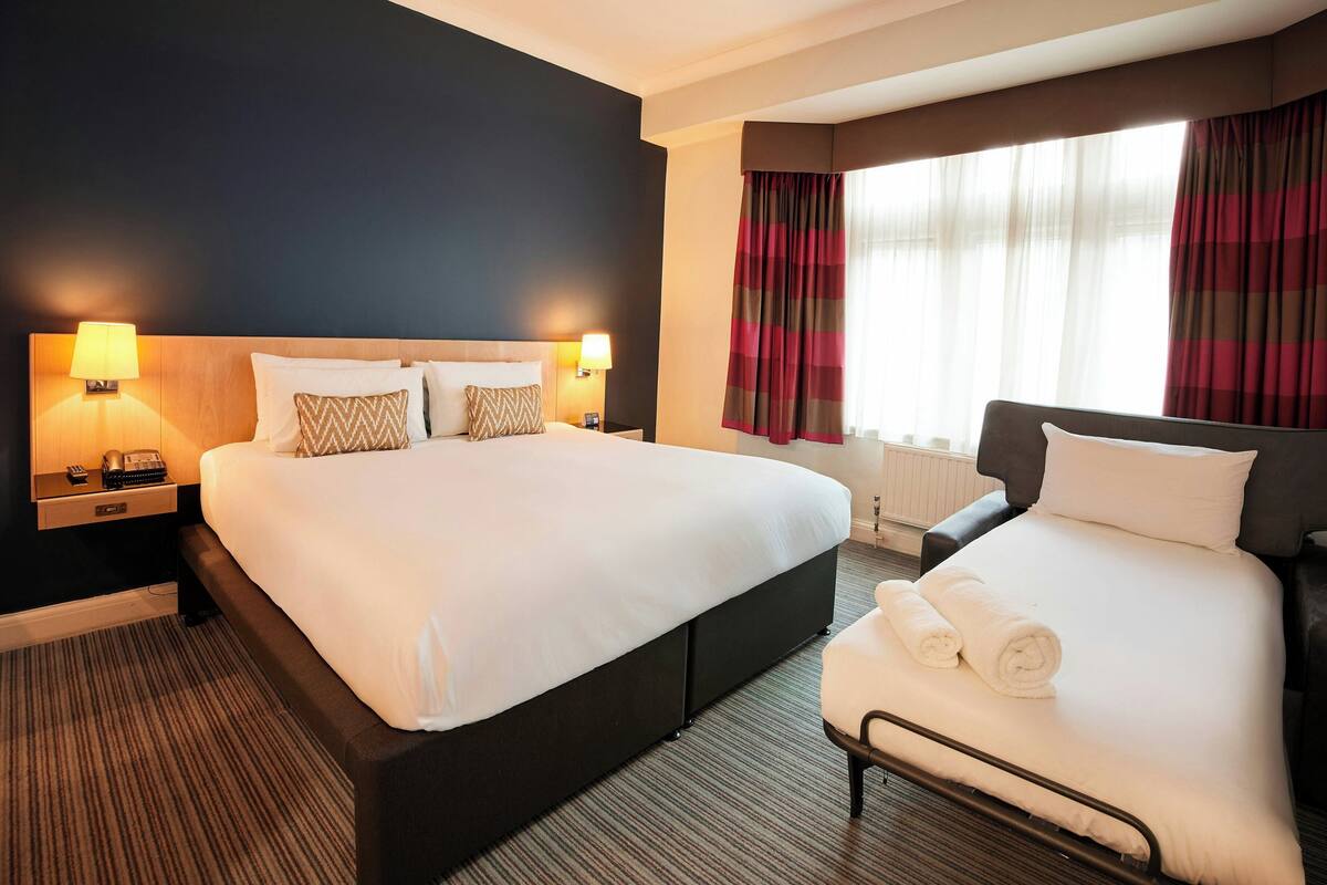 A hotel room with a large bed, a smaller bed, a nightstand with a lamp, and a window with curtains.