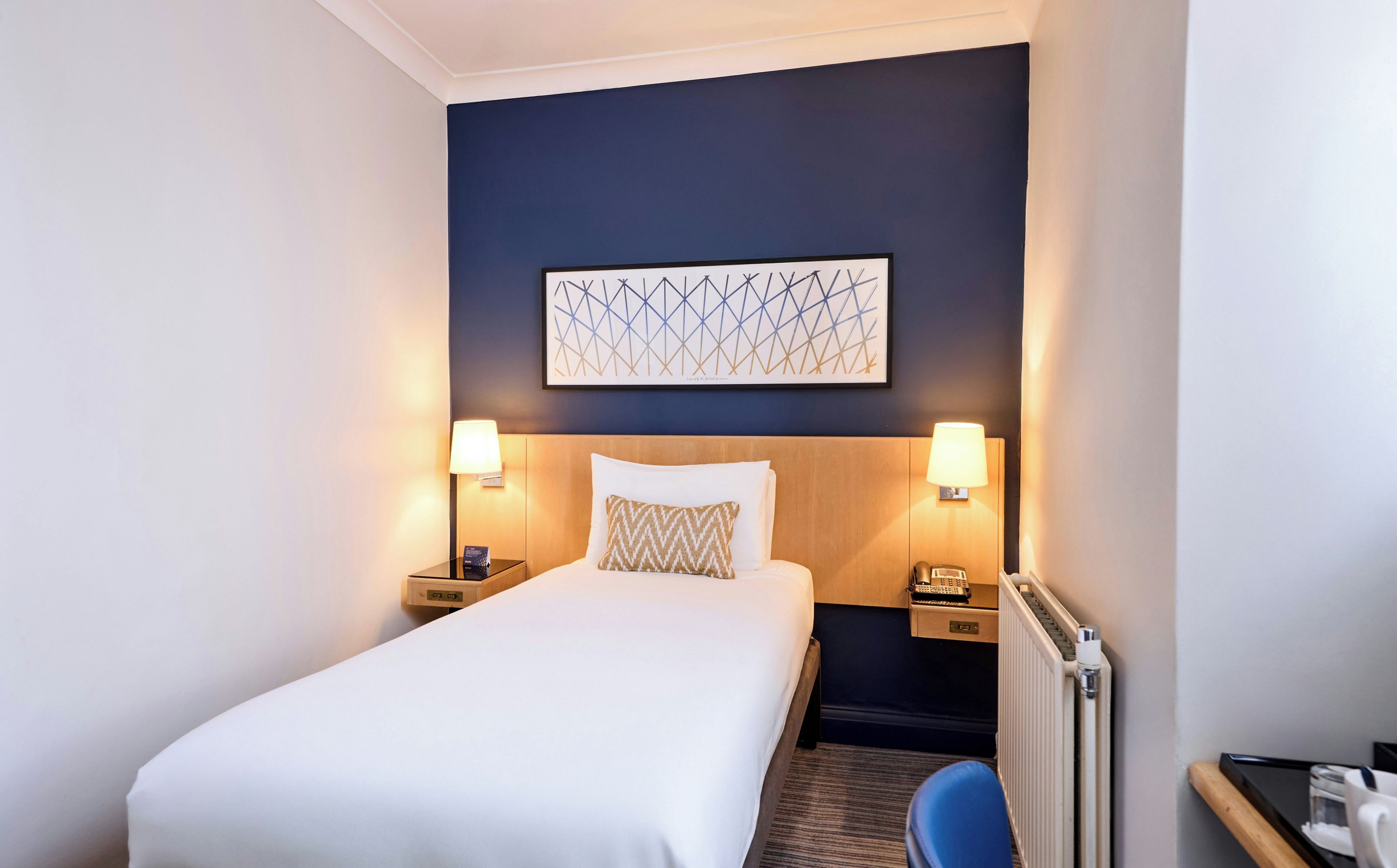 A hotel room with a bed, two bedside lamps, a nightstand, a wall art piece, and a radiator.