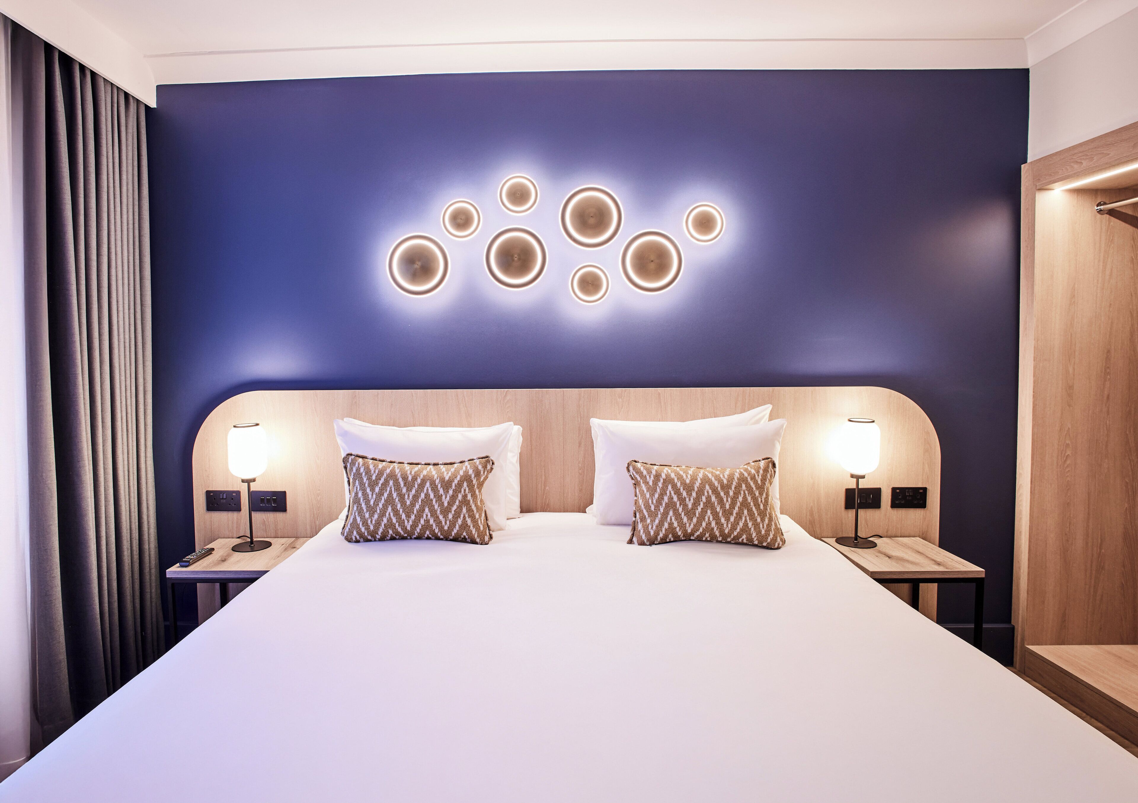Une chambre d’hôtel avec un grand lit, deux tables de chevet, un luminaire fixé au mur et une tête de lit en bois.
