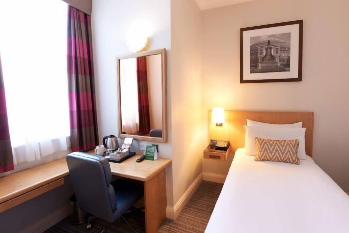 A hotel room with a bed, a desk, a chair, a lamp, a mirror, and a framed picture on the wall.