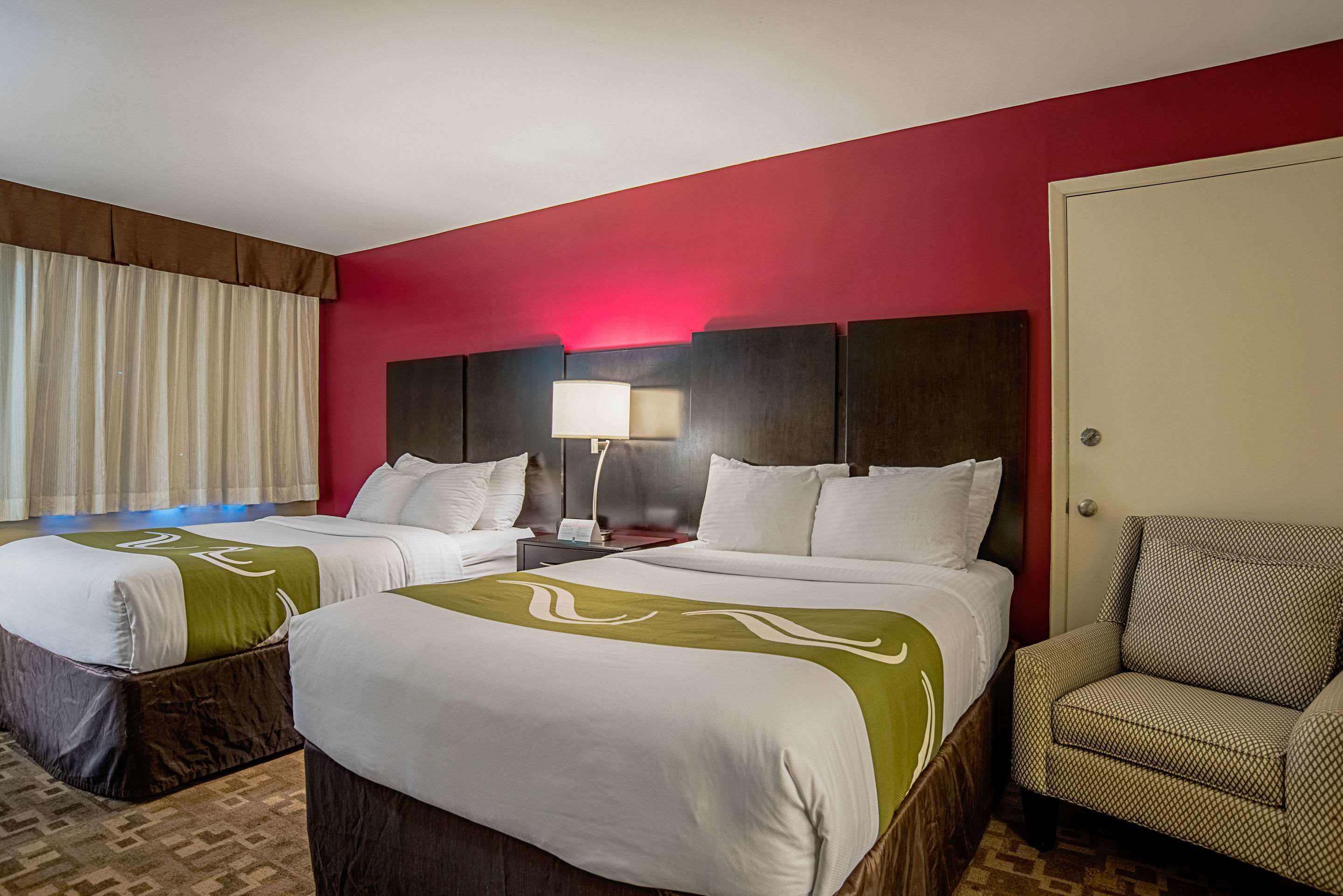 A hotel room with two beds, a chair, a nightstand with a lamp, and a red accent wall.