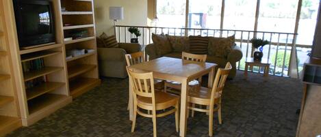 A dining area with a wooden table and chairs, a bookshelf, a television, and a large window with a view of the outdoors.