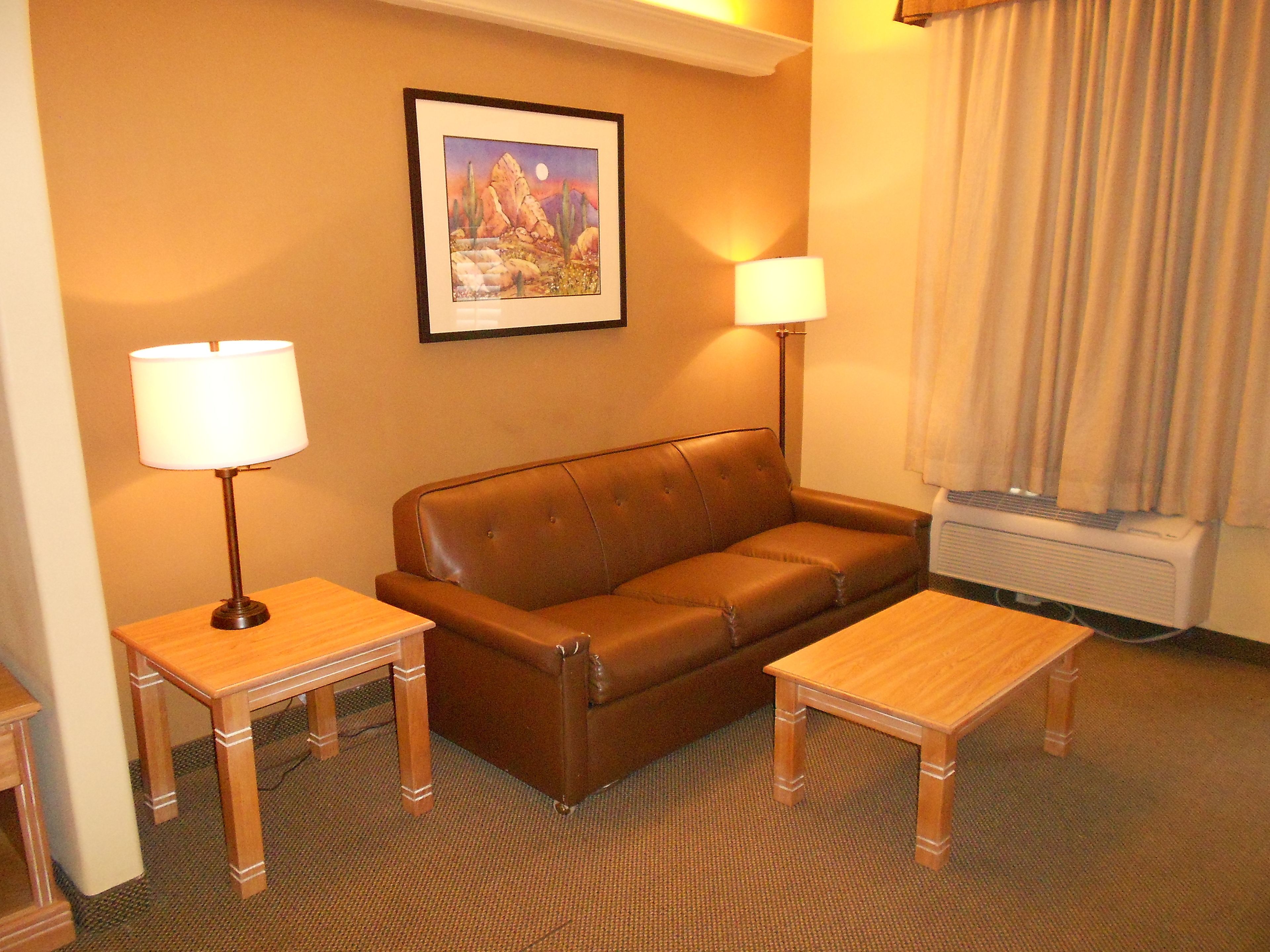 A hotel room with a brown leather sofa, two wooden side tables with lamps, a framed picture on the wall, and a window with curtains.