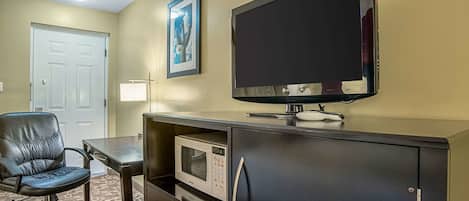 Et hotelværelse med et fladskærms-tv på væggen, et skrivebord med mikroovn, en sort læderstol og en dør.