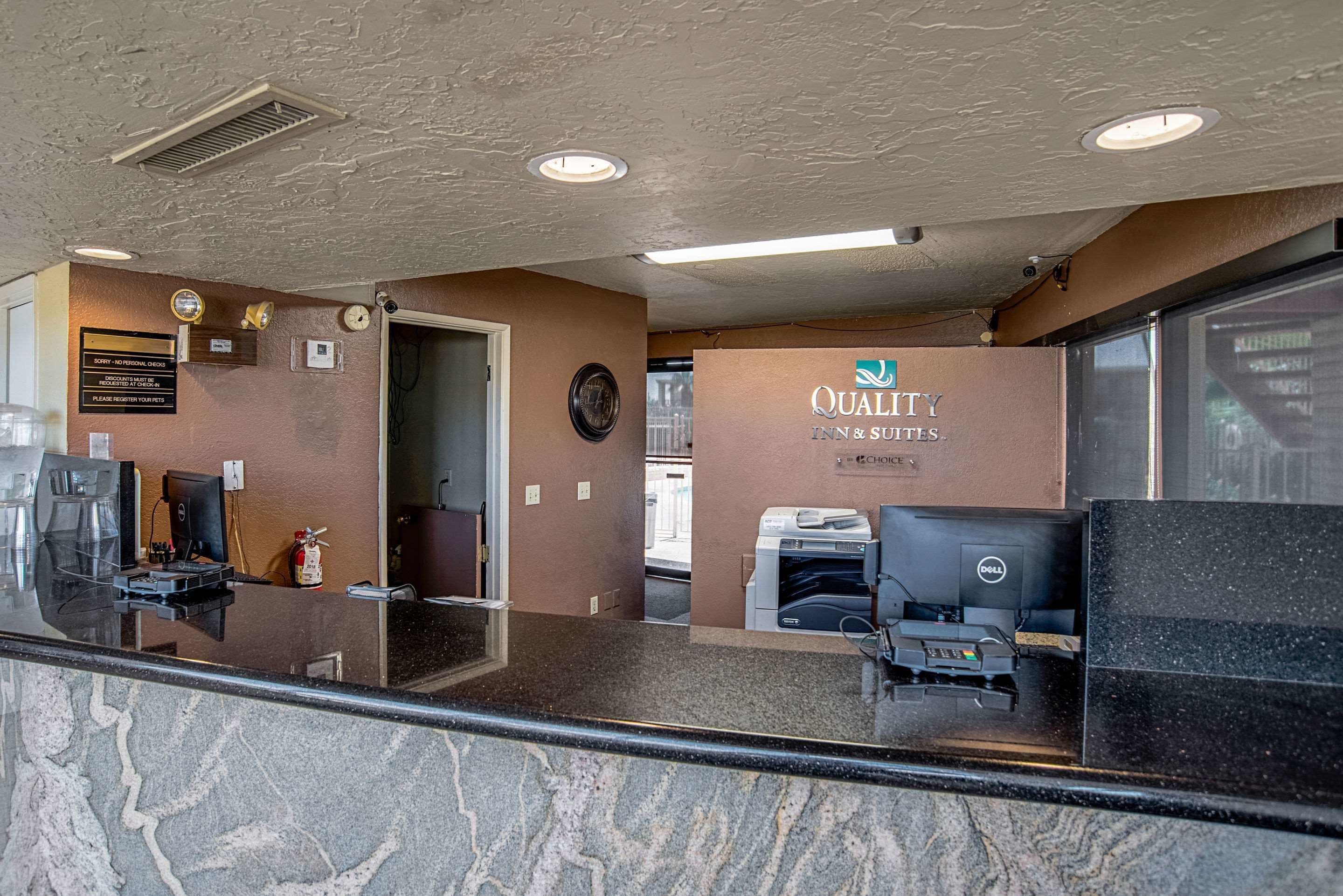 En receptionsdisk med et skilt for Quality Inn & Suites, en kaffemaskine, en brandslukker og en Dell-printer.