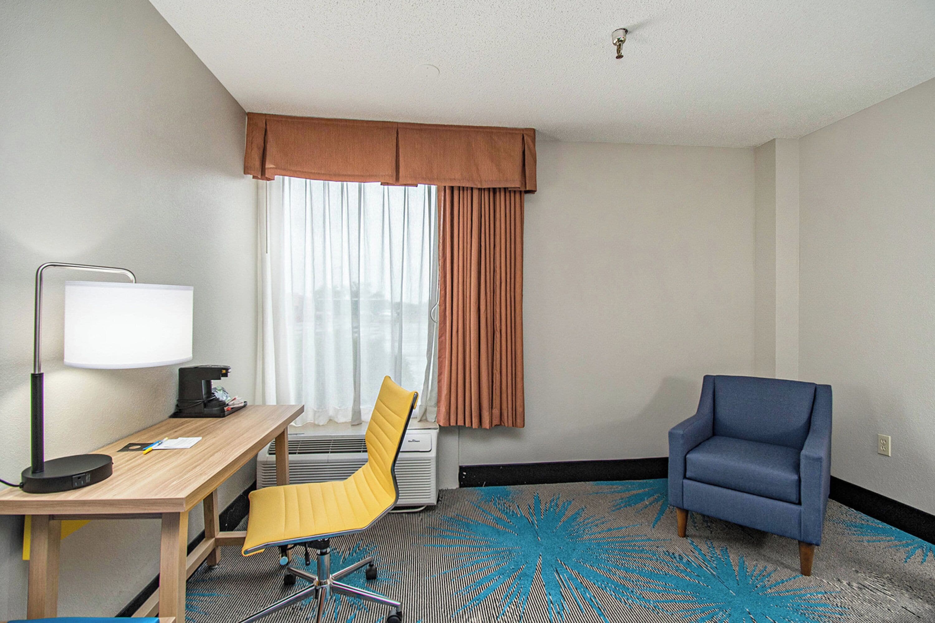 A hotel room with a desk, chair, lamp, and a blue armchair.