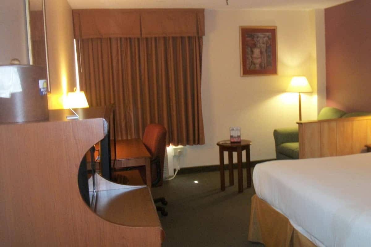 A hotel room with a bed, a desk, a chair, a small table, a lamp, and a picture on the wall.