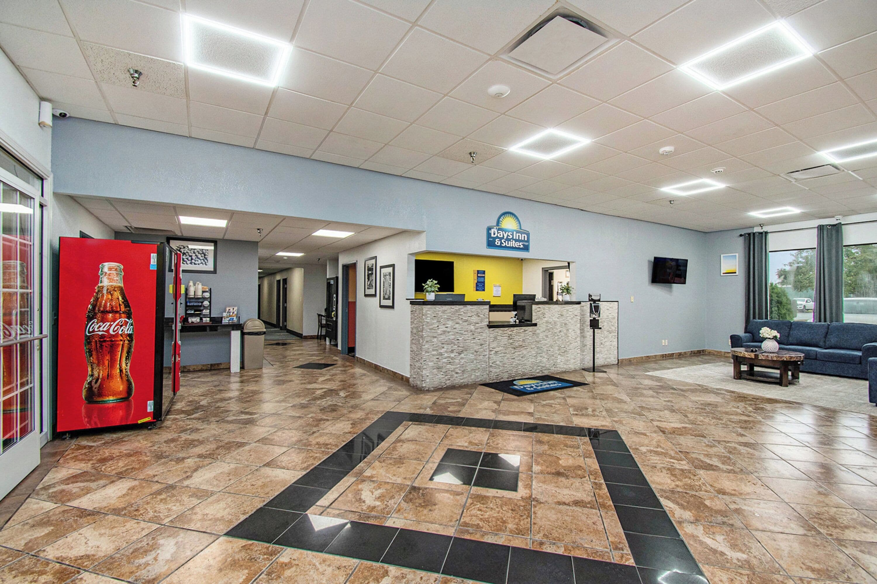 A hotel lobby with a reception desk, a Coca-Cola refrigerator, a large window, and a seating area.