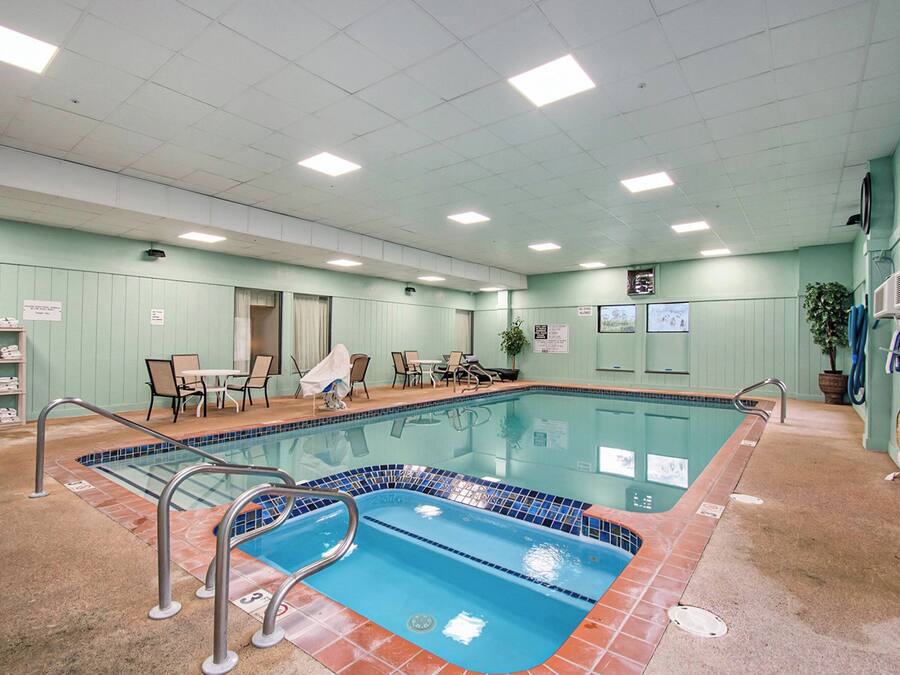 An indoor swimming pool with a tiled edge, a lounge area with chairs and a table, and a ceiling with recessed lighting.