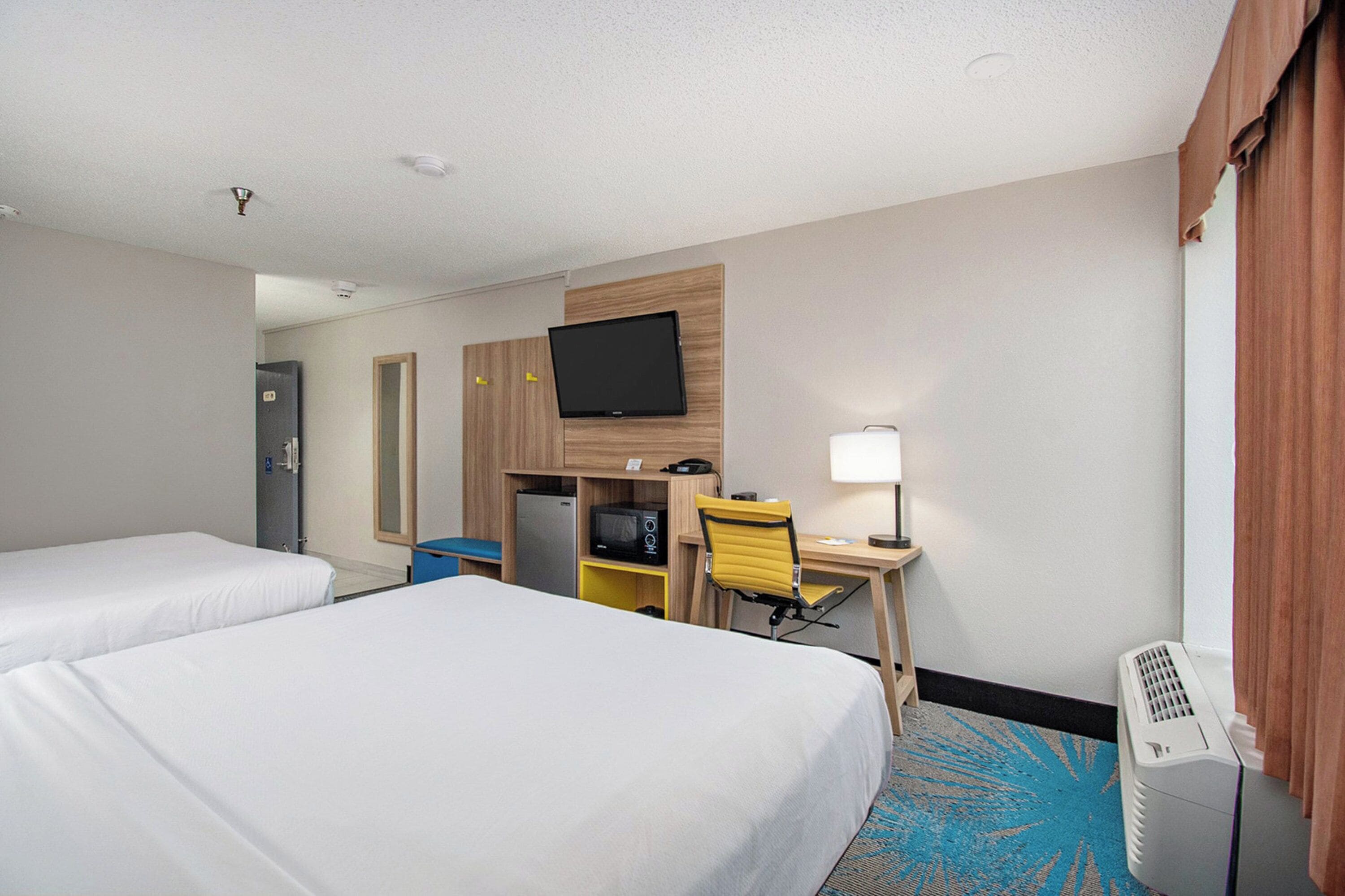 A hotel room with two beds, a desk with a chair, a television mounted on the wall, a desk lamp, and a heater.