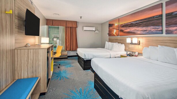 A hotel room with two beds, a desk, a chair, a TV, an air conditioning unit, and a large sunset painting on the wall.