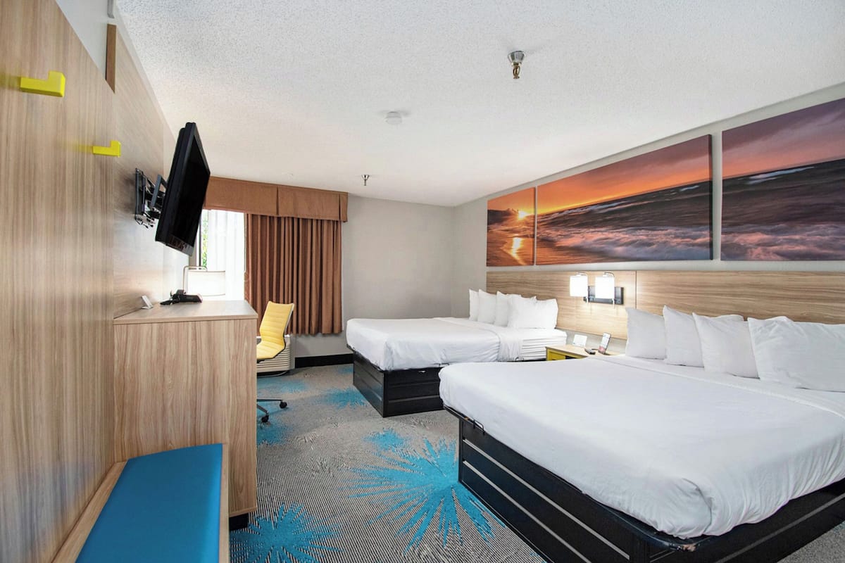 A hotel room with two beds, a desk, a TV, and a large sunset painting on the wall.