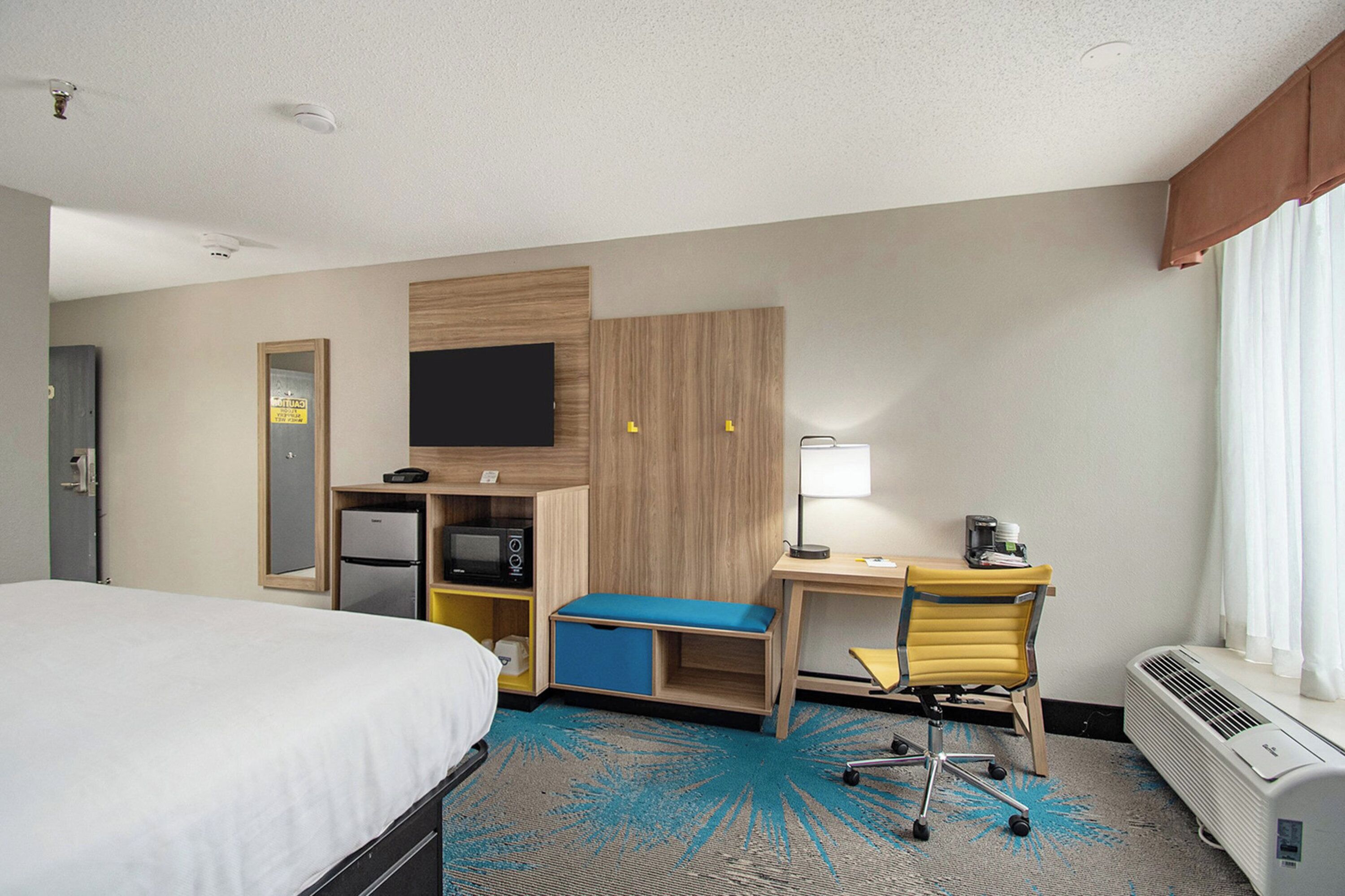 A hotel room with a bed, desk, chair, TV, mini-fridge, and a window with curtains.