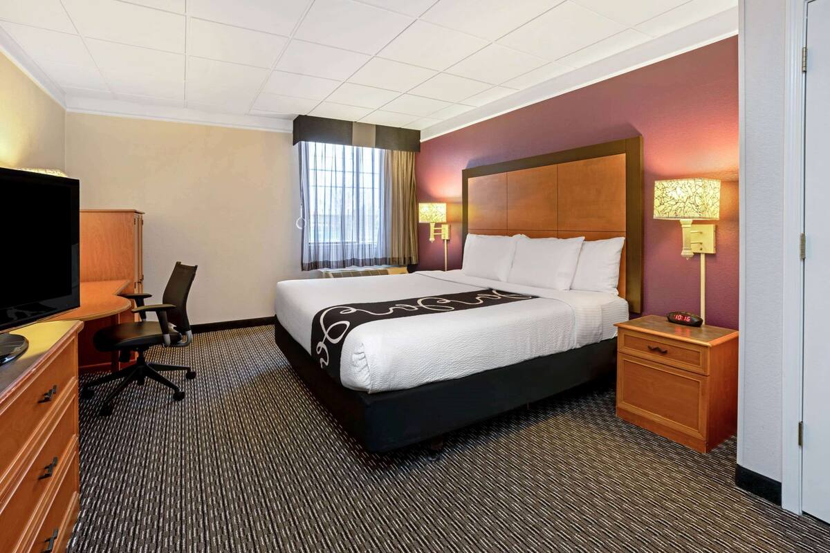 Quarto de hotel com cama grande, escrivaninha com televisão, poltrona, mesa de cabeceira com abajur e janela com cortinas.
