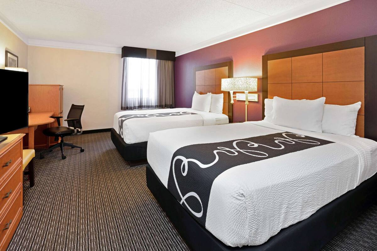 Quarto de hotel com duas camas, uma escrivaninha, uma cadeira, televisão e janela com cortinas.