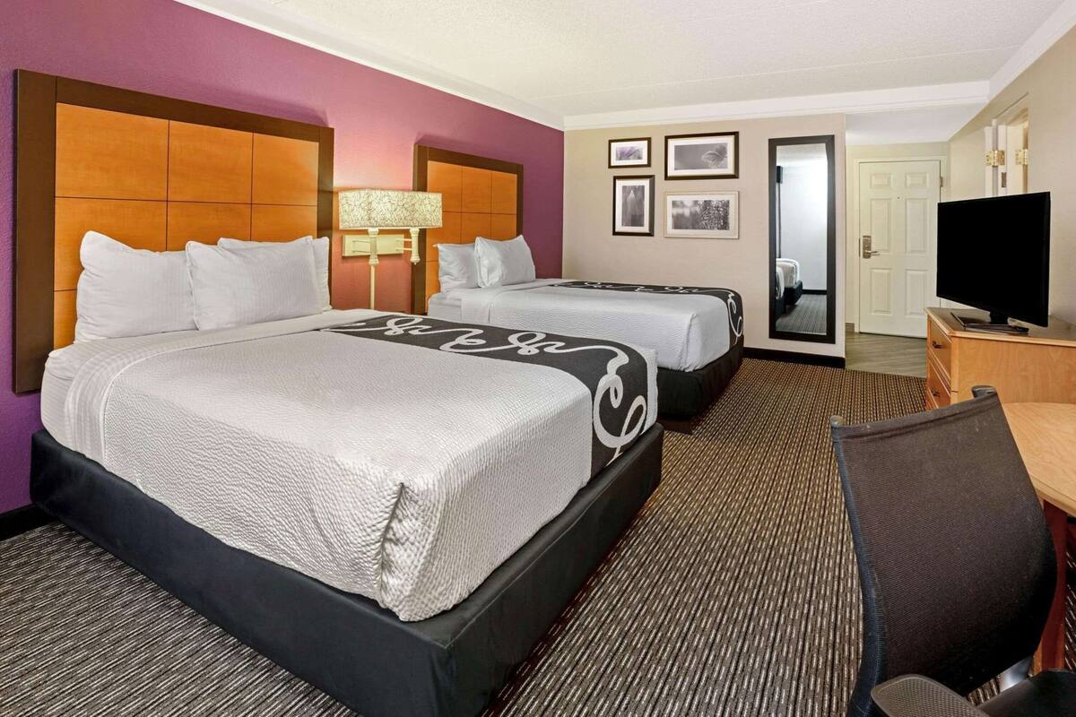 Quarto de hotel com uma cama grande, duas camas menores, uma escrivaninha, uma cadeira, televisão e quadros nas paredes.