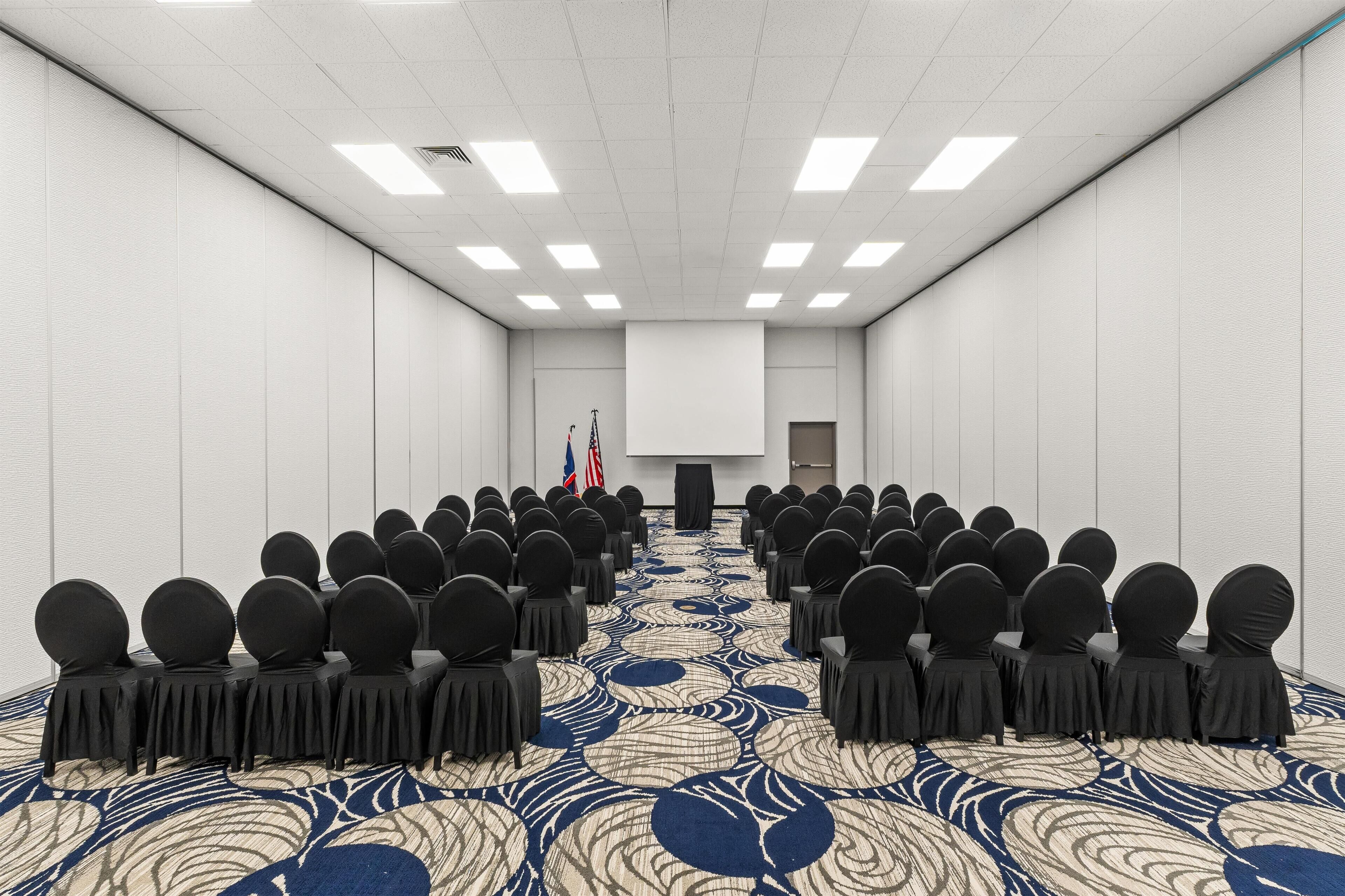 A conference room with rows of chairs facing a presentation screen, flags, and a patterned carpet.
